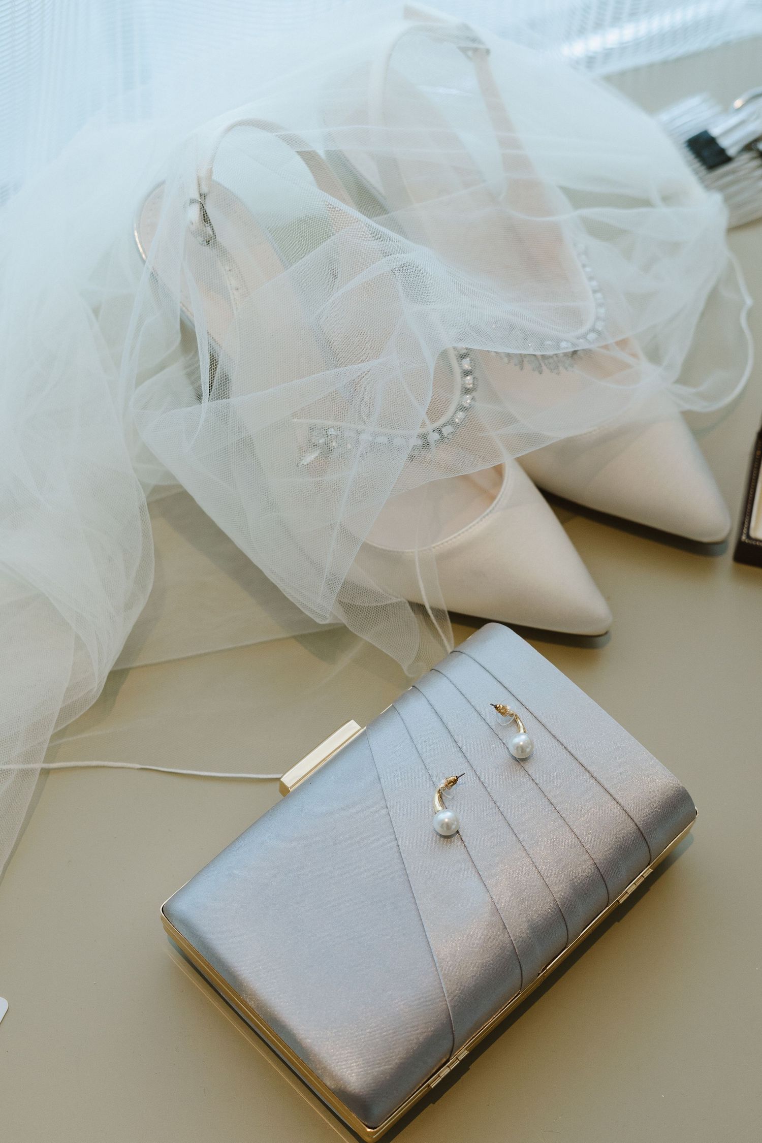 White bridal shoes with tulle details and silver clutch bag arranged on light surface.