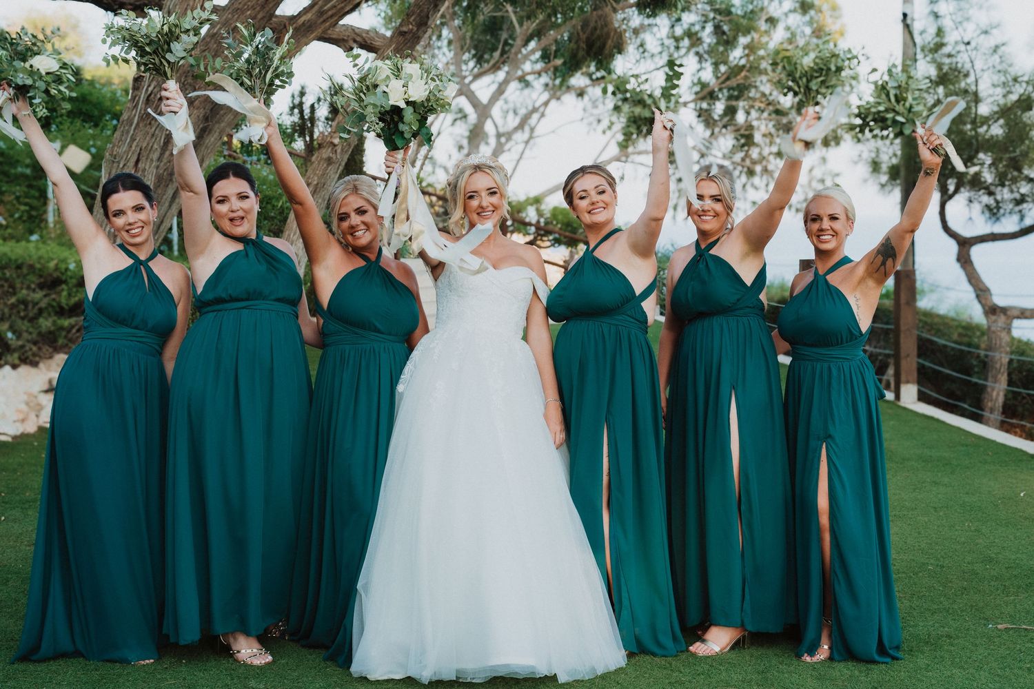 Wedding party in emerald green dresses poses joyfully with bride in white gown on manicured lawn.