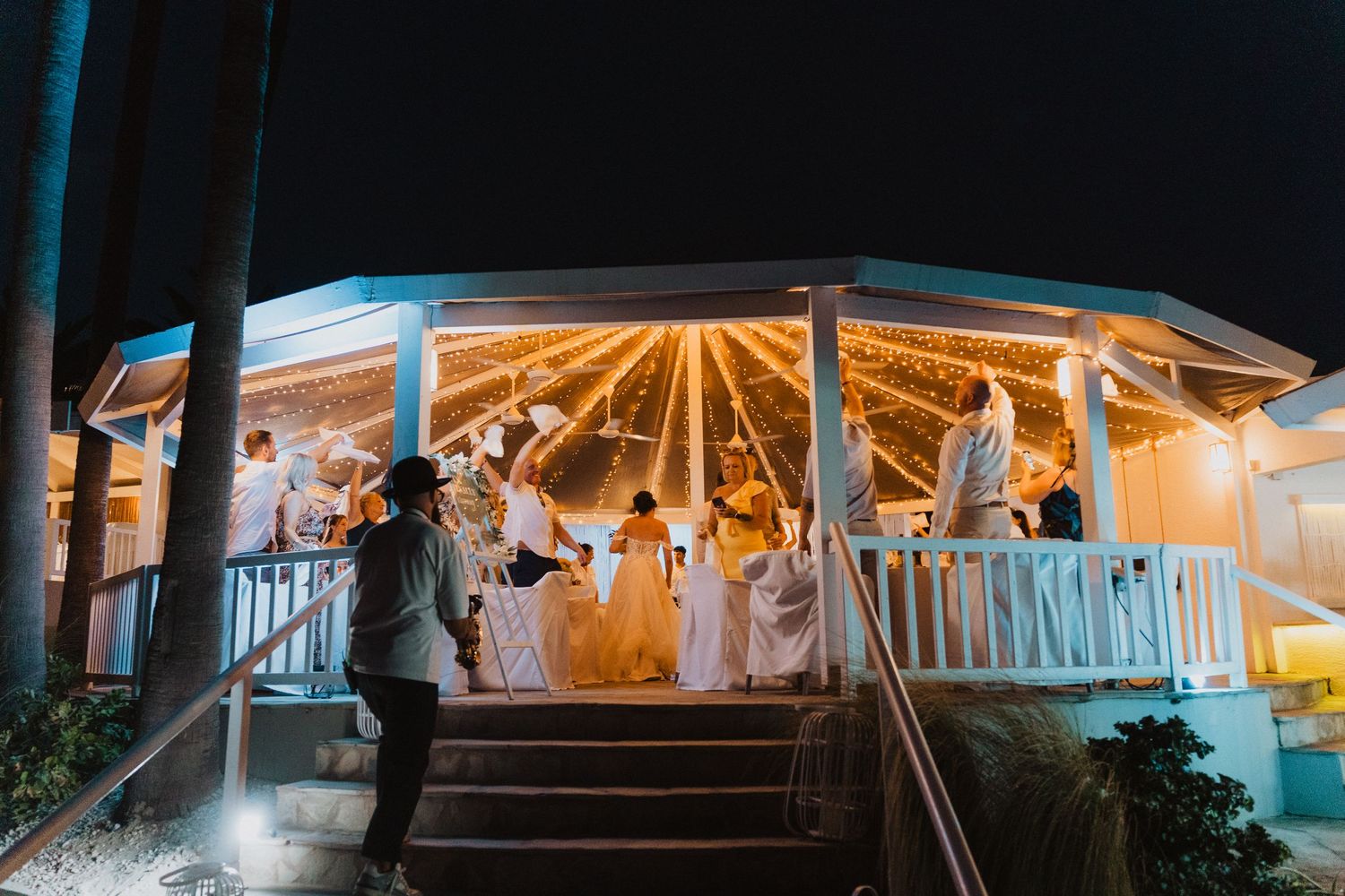 People dancing and celebrating at night in an illuminated white gazebo decorated with warm glowing lights.