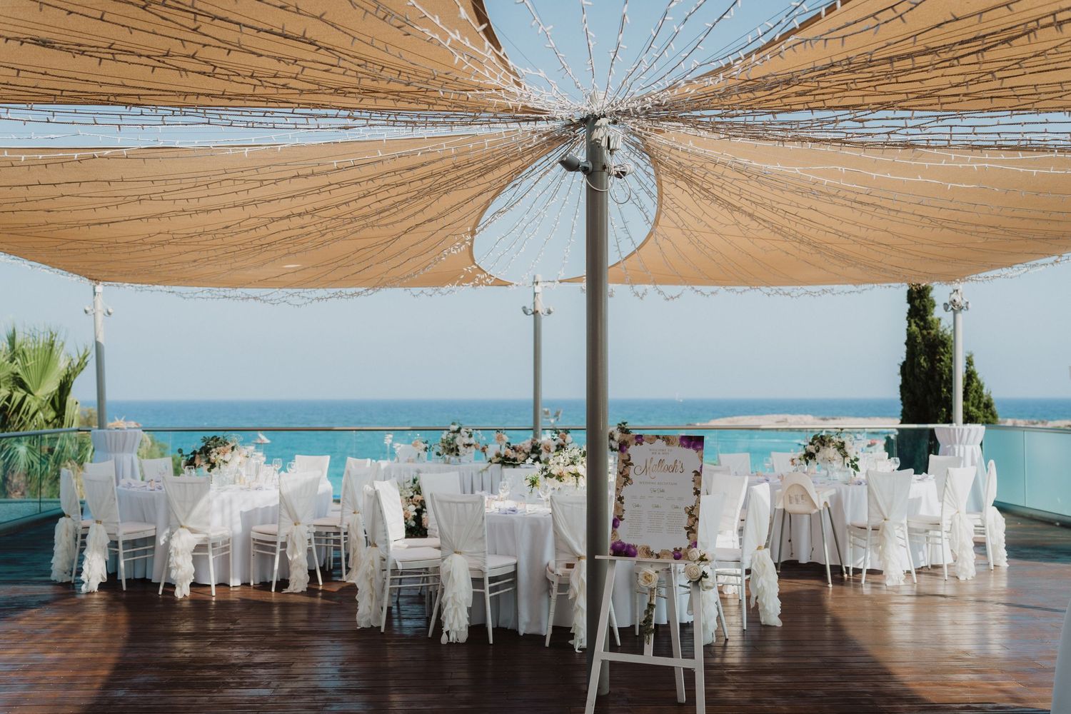 Outdoor wedding reception setup with white chairs under beige canopy overlooking the ocean.