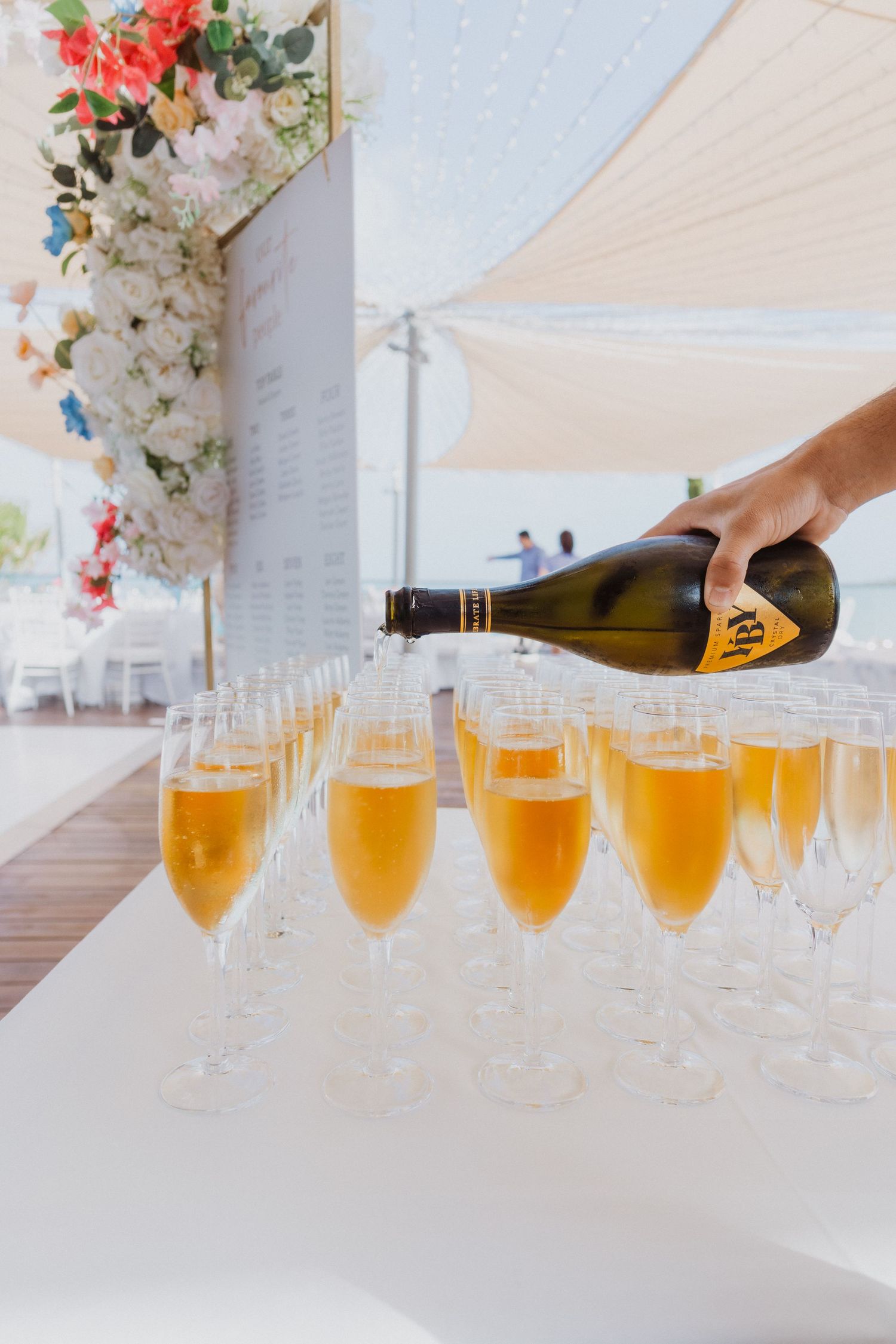 Champagne being poured into multiple glasses at an elegant event with floral decorations and white umbrellas.