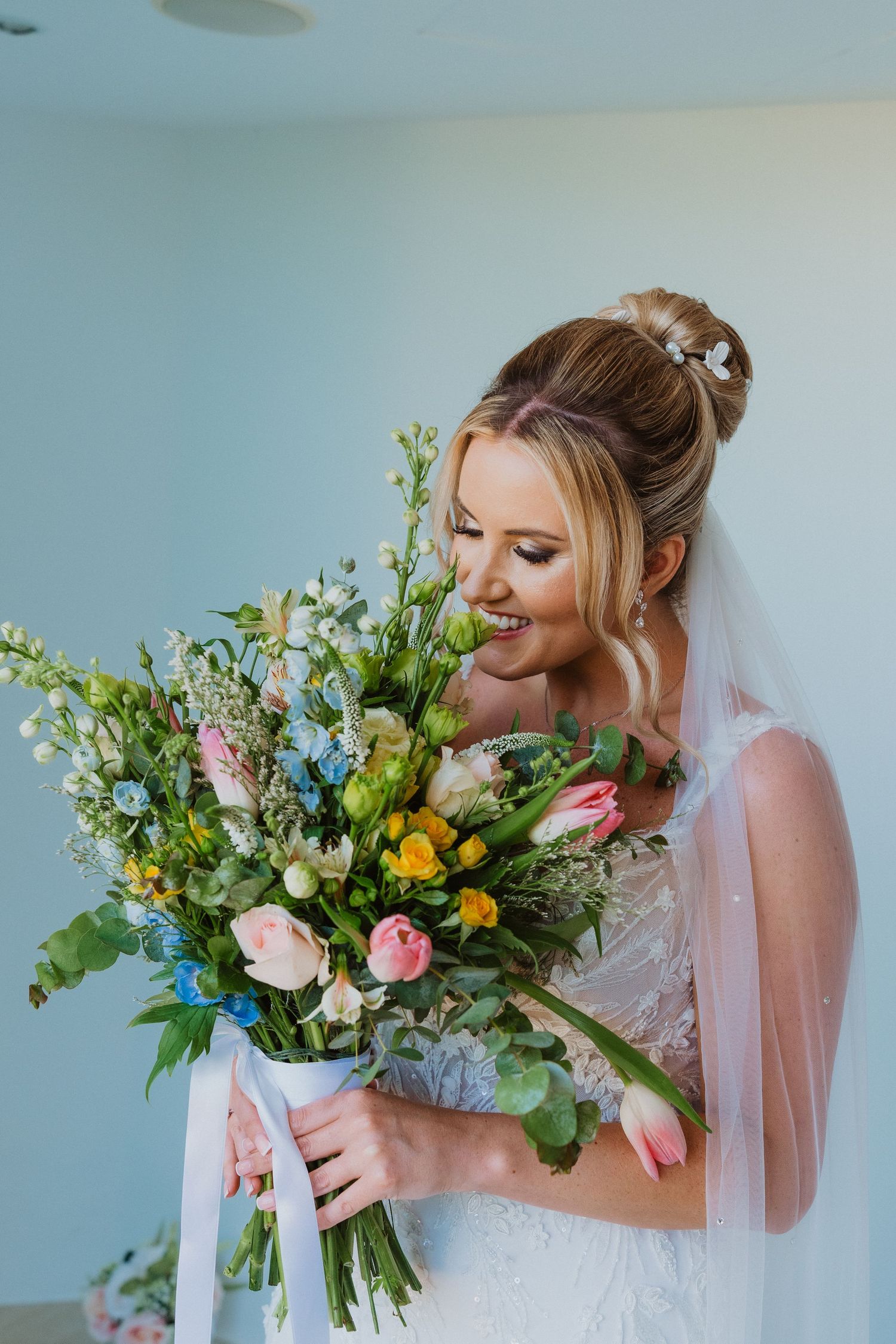 A bride in a white wedding dress holds a vibrant spring bouquet with yellow and pink flowers against a light blue wall.