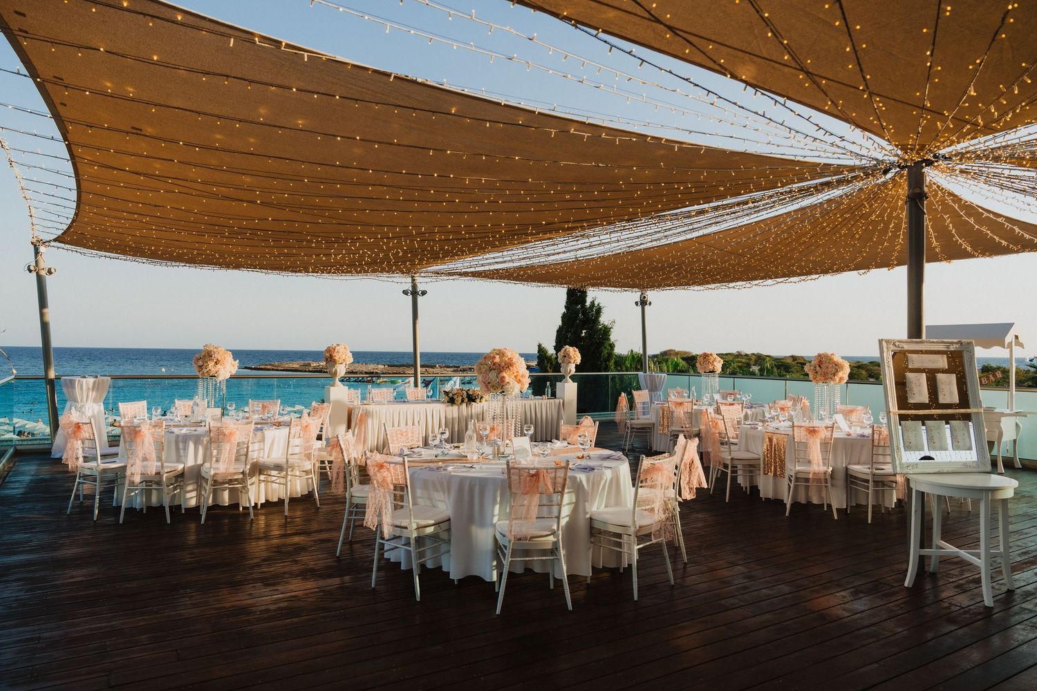 Elegant outdoor wedding reception setup with white chairs under beige sail shades overlooking the ocean.
