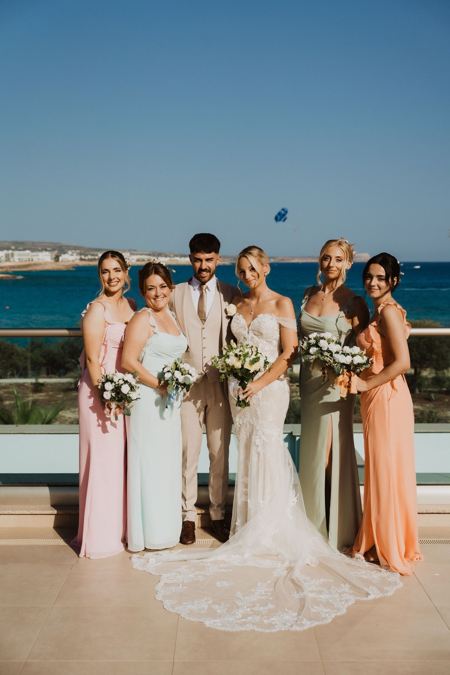 Bridal party in pastel dresses poses against an oceanfront backdrop.