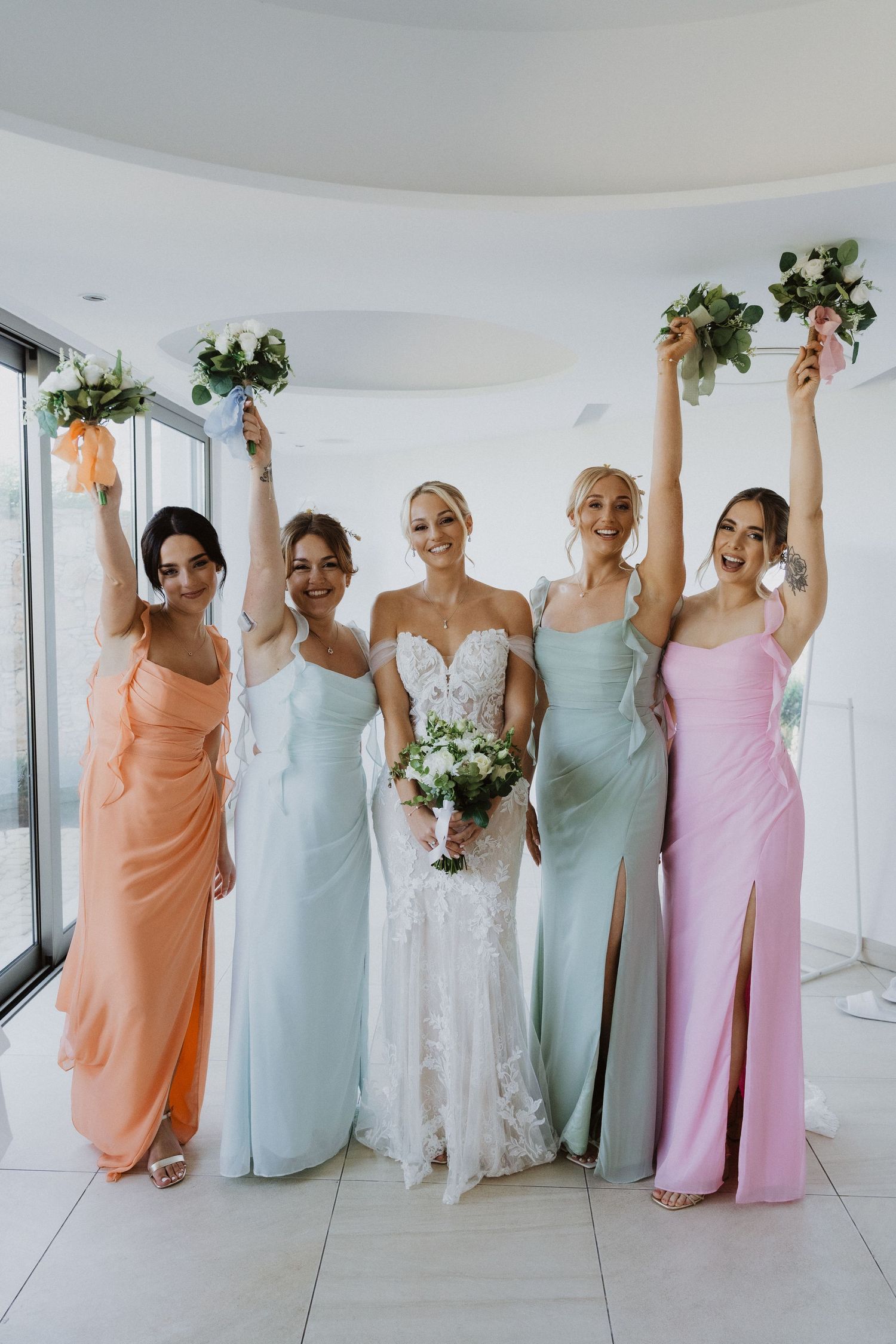 Bridesmaids in pastel dresses celebrate joyfully while holding bouquets in a bright venue.