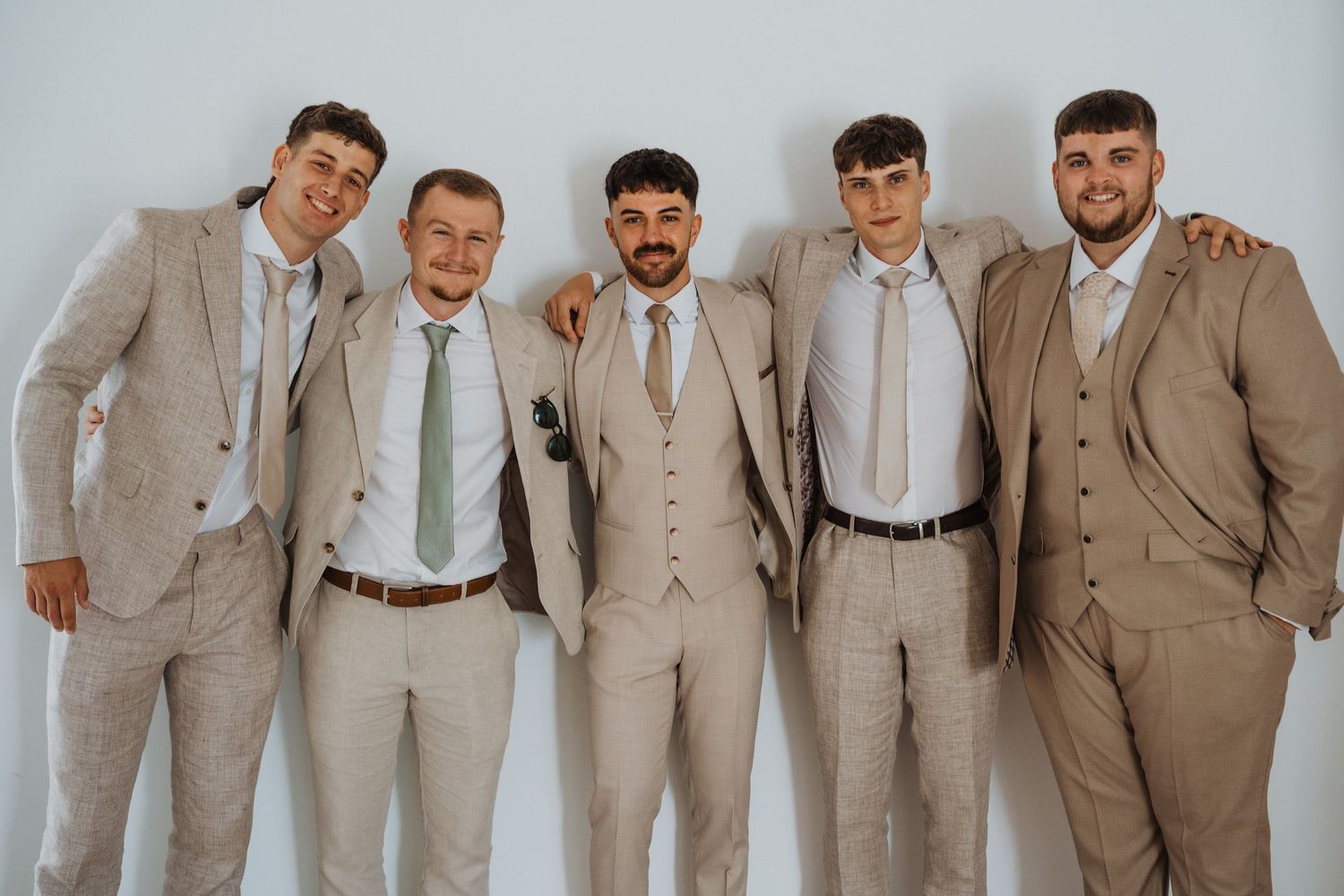 Groomsmen in matching beige suits posing together against a white wall.