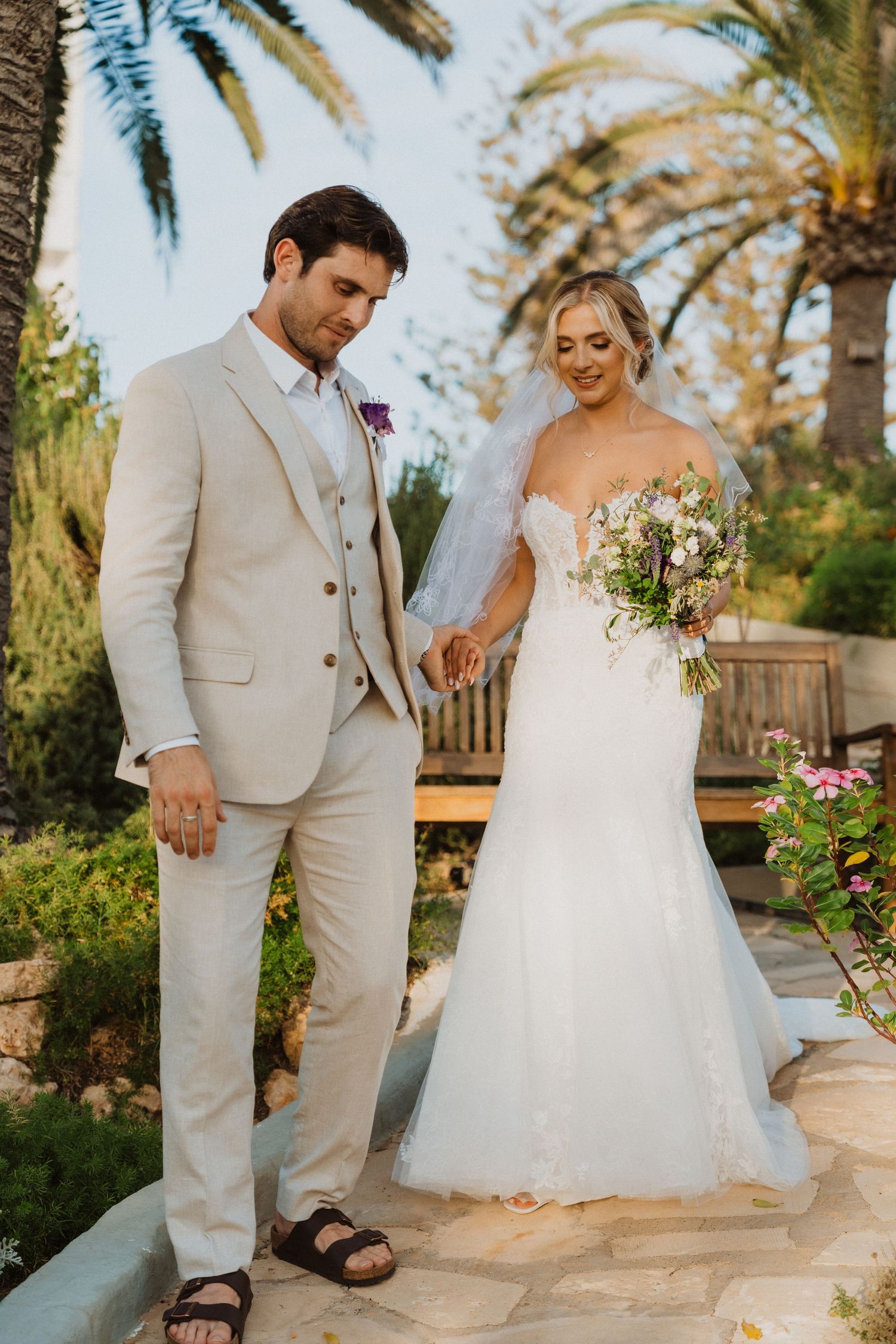 A wedding couple poses in a tropical garden setting with palm trees and flowers.