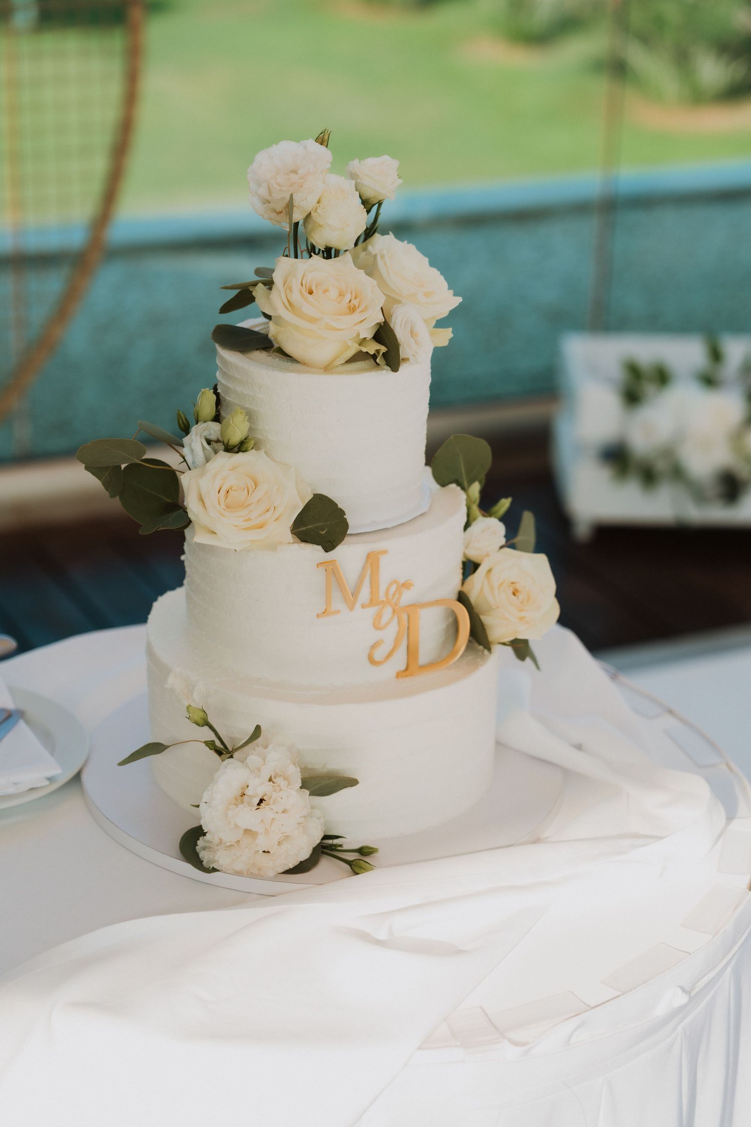 Elegant three-tier white wedding cake with cream roses and gold Mr & D monogram topper on a white draped table.