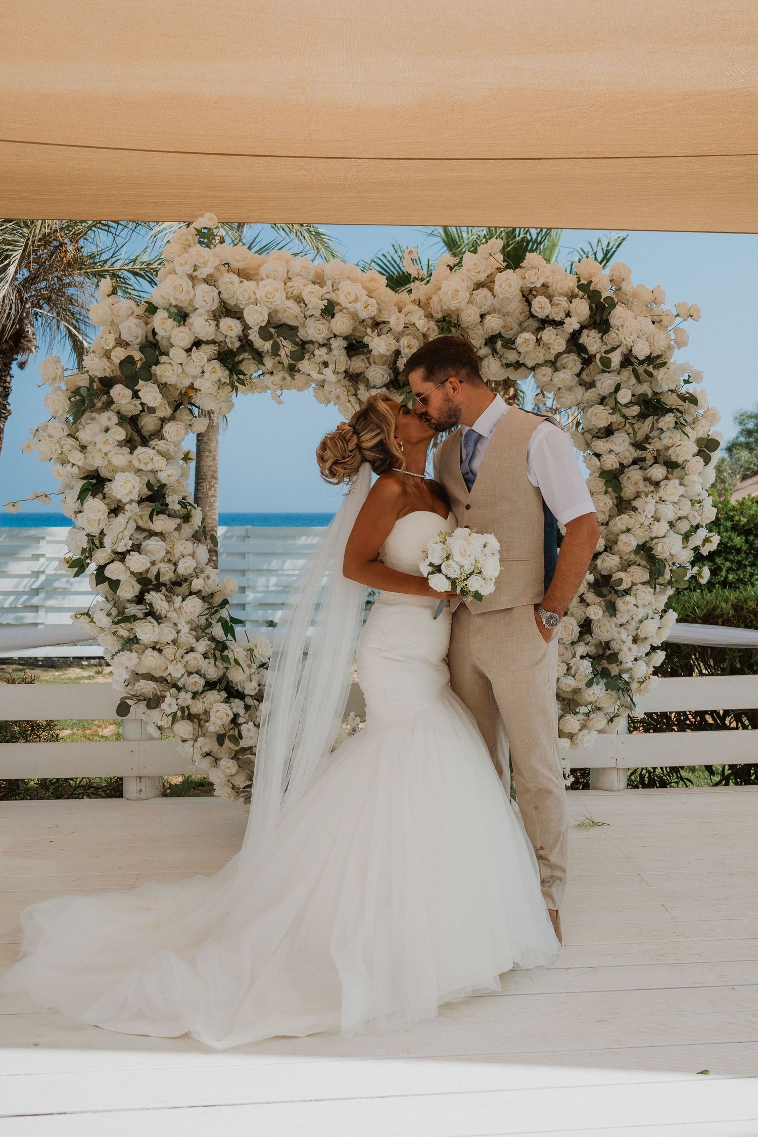 Bride and groom kissing under a white floral arch at a beachside wedding ceremony.