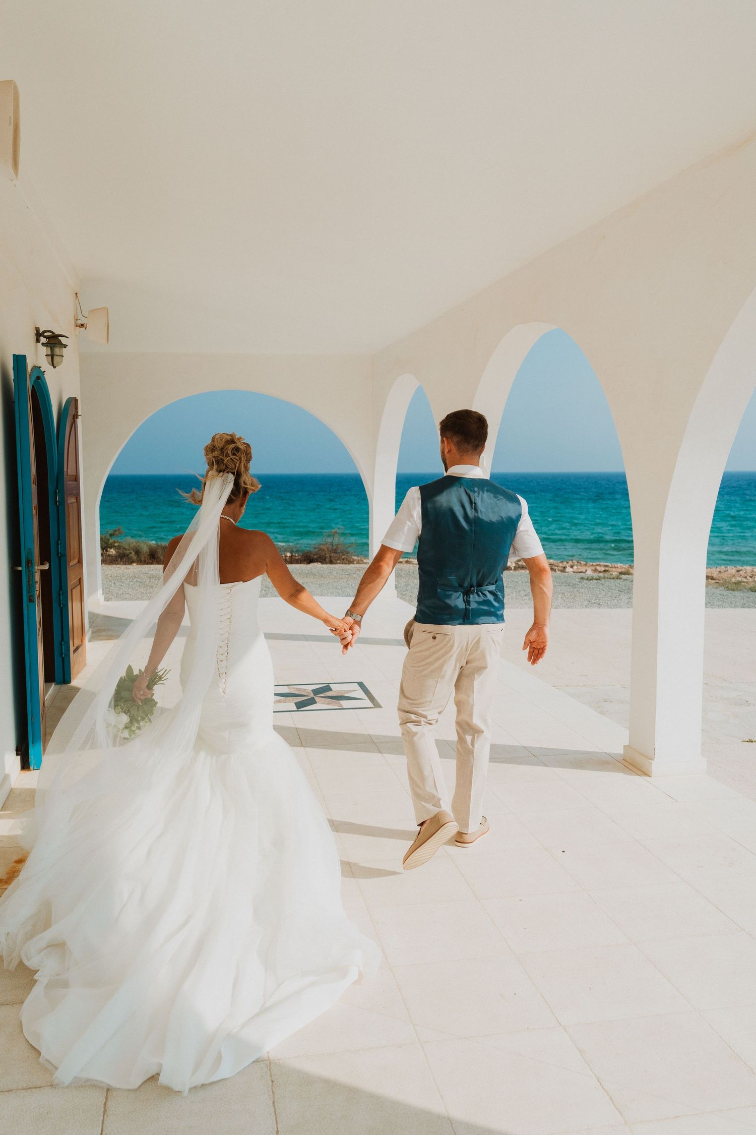 Bride and groom holding hands walking through white arched colonnade toward turquoise sea at Mediterranean wedding venue.