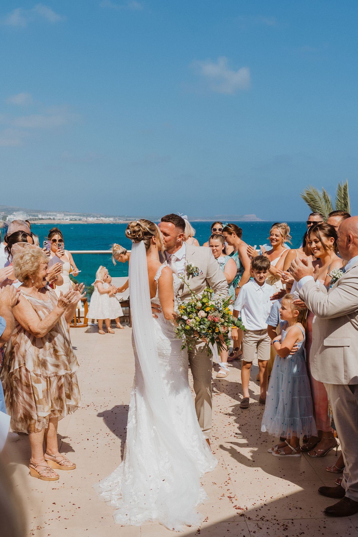 Wedding guests on a sunny beach watching a couple's first kiss during a seaside ceremony.