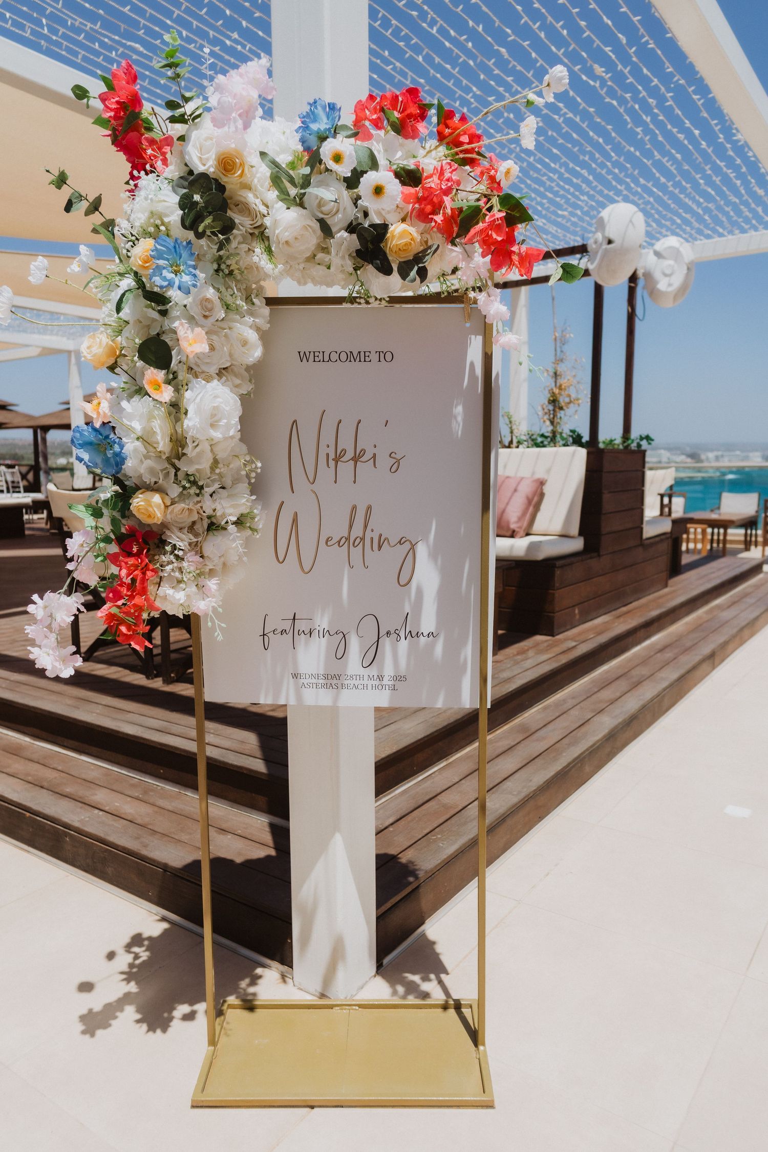 A wedding welcome sign with floral decorations stands on a gold frame at a beachfront venue with ocean views.