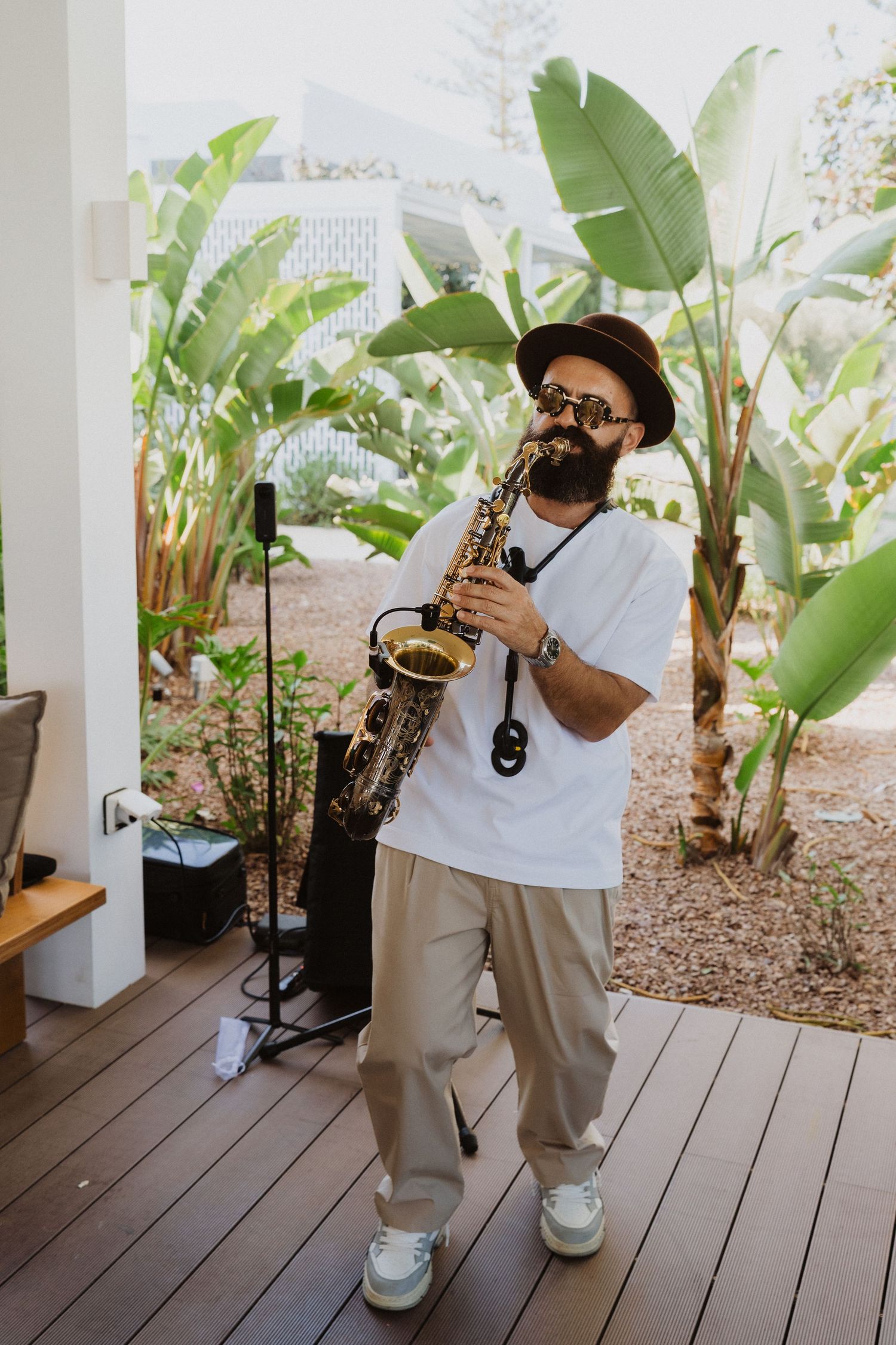 A musician plays saxophone on an outdoor deck surrounded by tropical plants and banana leaves.