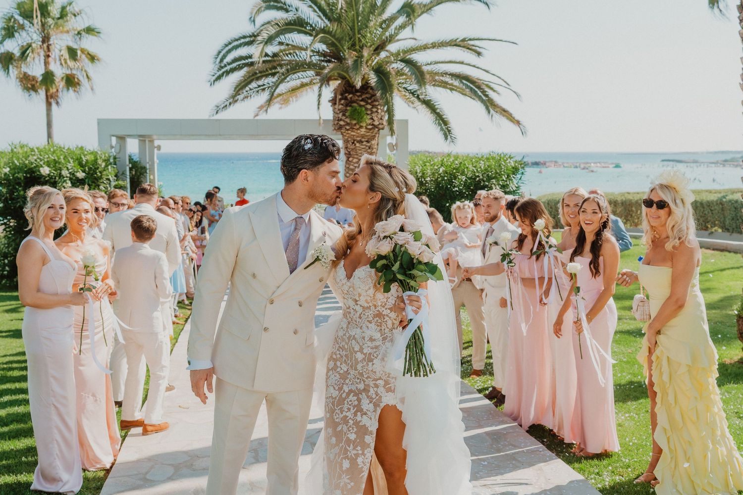 A wedding celebration takes place on a sunny lawn with palm trees and ocean views, surrounded by guests in pastel attire.
