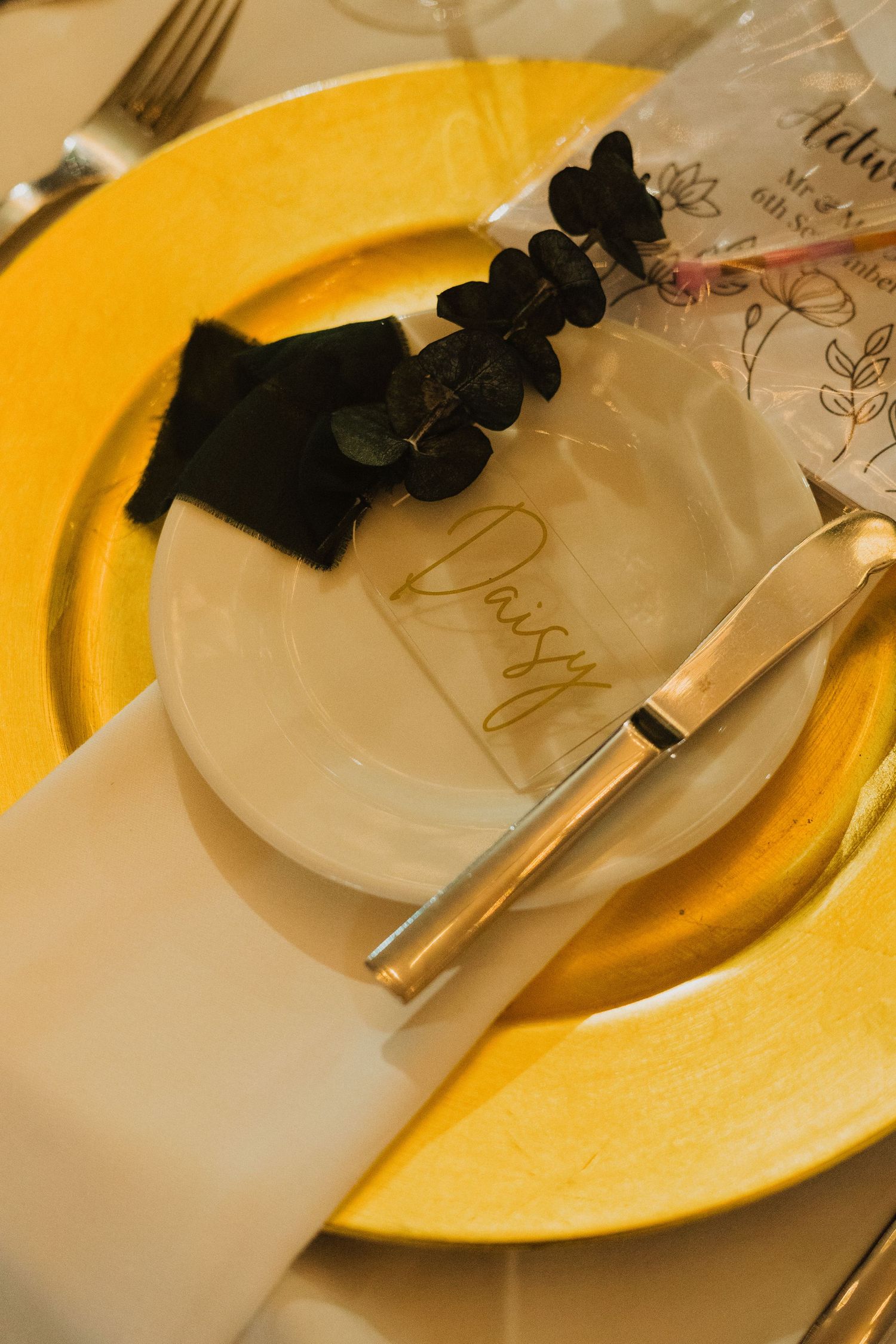 A formal place setting features a yellow charger plate, white napkin, and black floral decoration.