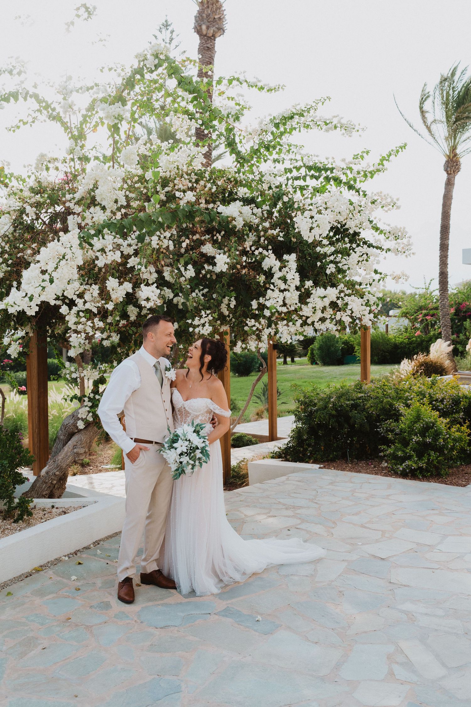 A couple poses under a blooming white bougainvillea archway on a stone patio during a tropical destination wedding.