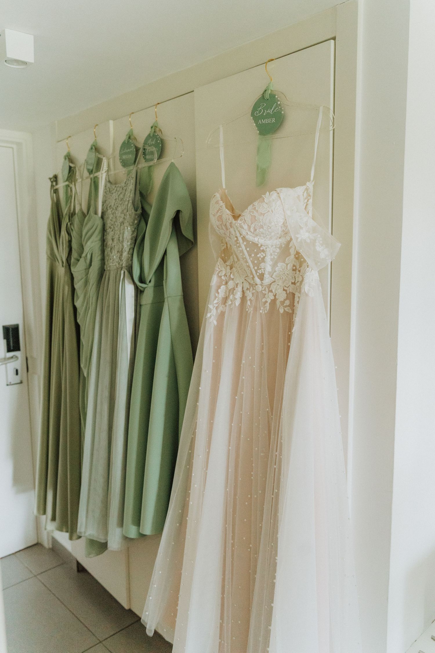 Elegant sage green bridesmaid dresses and a blush pink wedding gown hanging on matching hangers against a cream wall.
