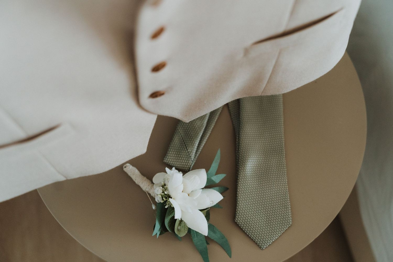 Close up of a sage green tie with white floral boutonniere against a light colored suit jacket.