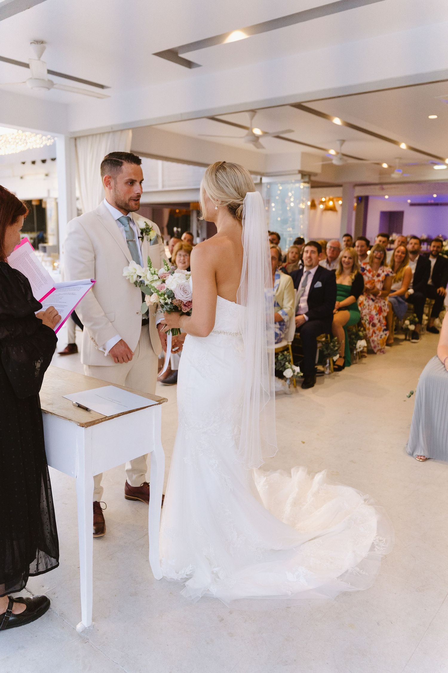 A wedding ceremony takes place in an elegant indoor venue with white decor and purple lighting accents.