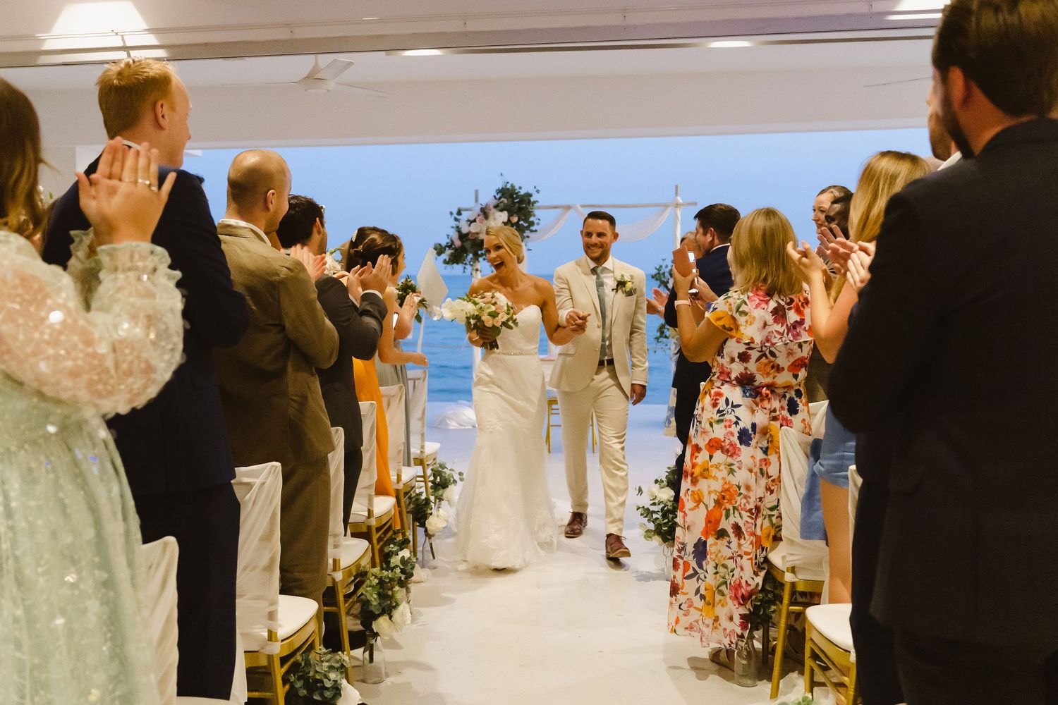 Wedding guests applaud as the newlyweds walk down the aisle in an indoor ceremony with ocean views.