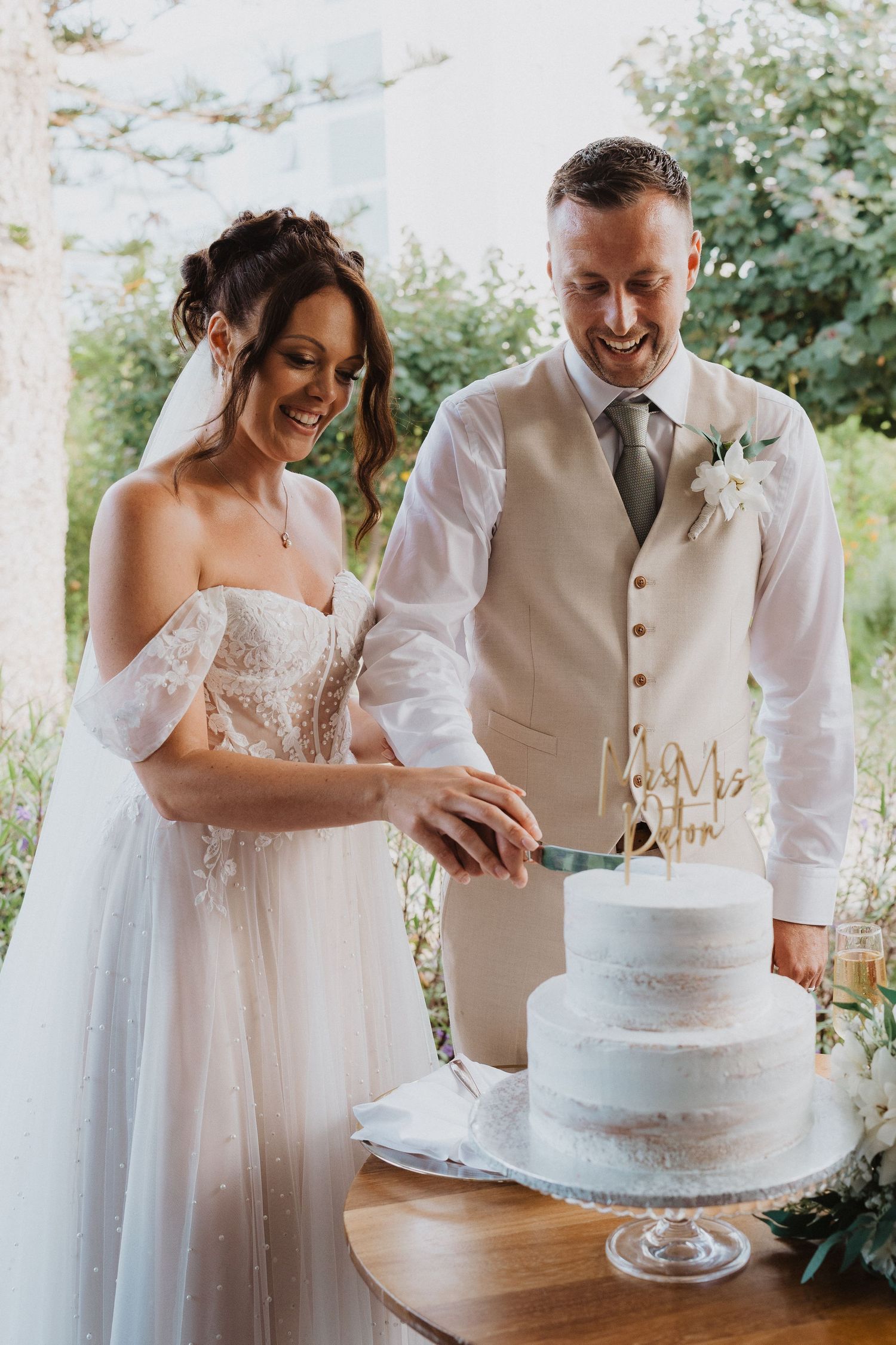 A newlywed couple smiles as they cut into their elegant white semi-naked wedding cake on a glass cake stand.