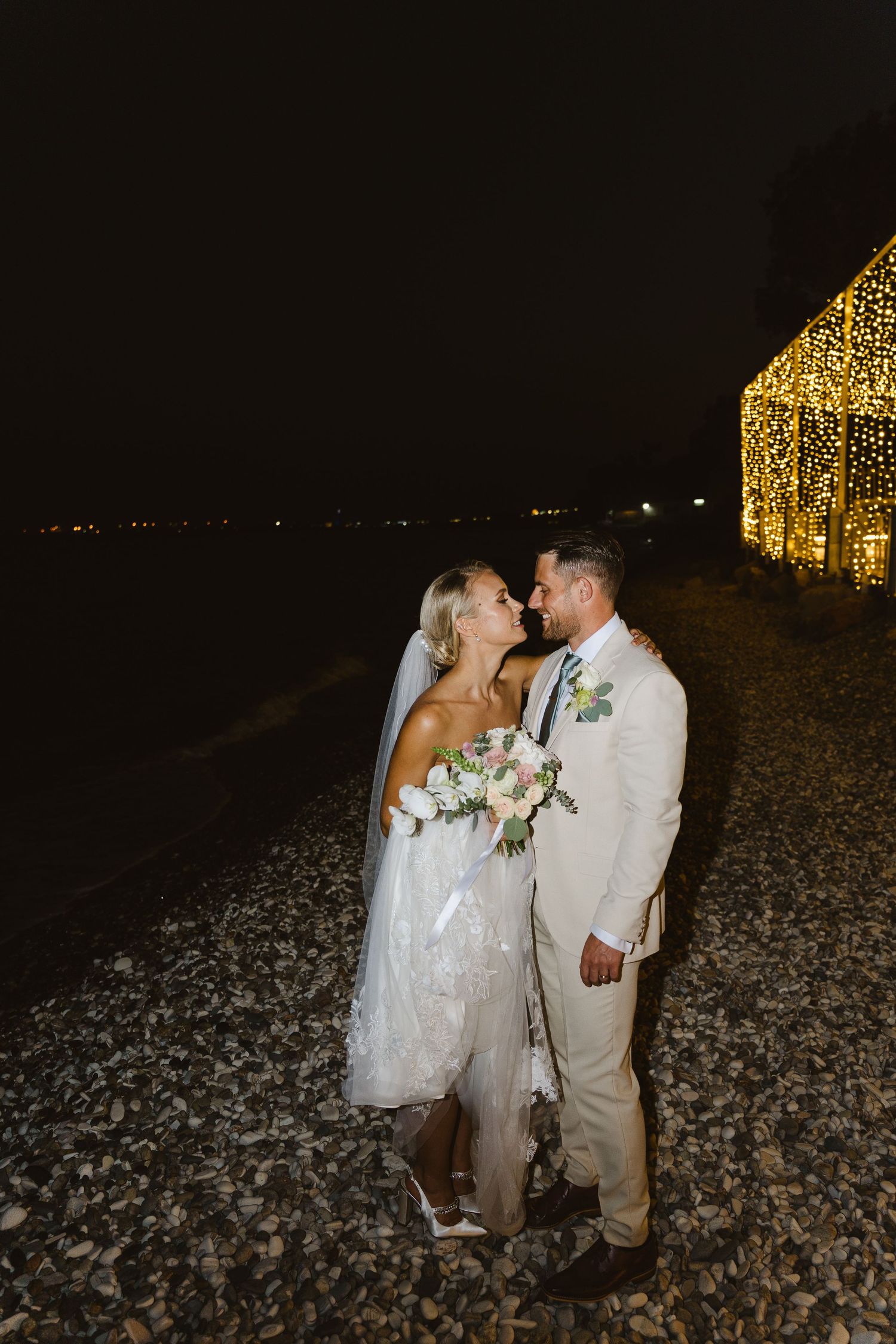 Romantic wedding portrait with glowing structure and ocean backdrop at night.