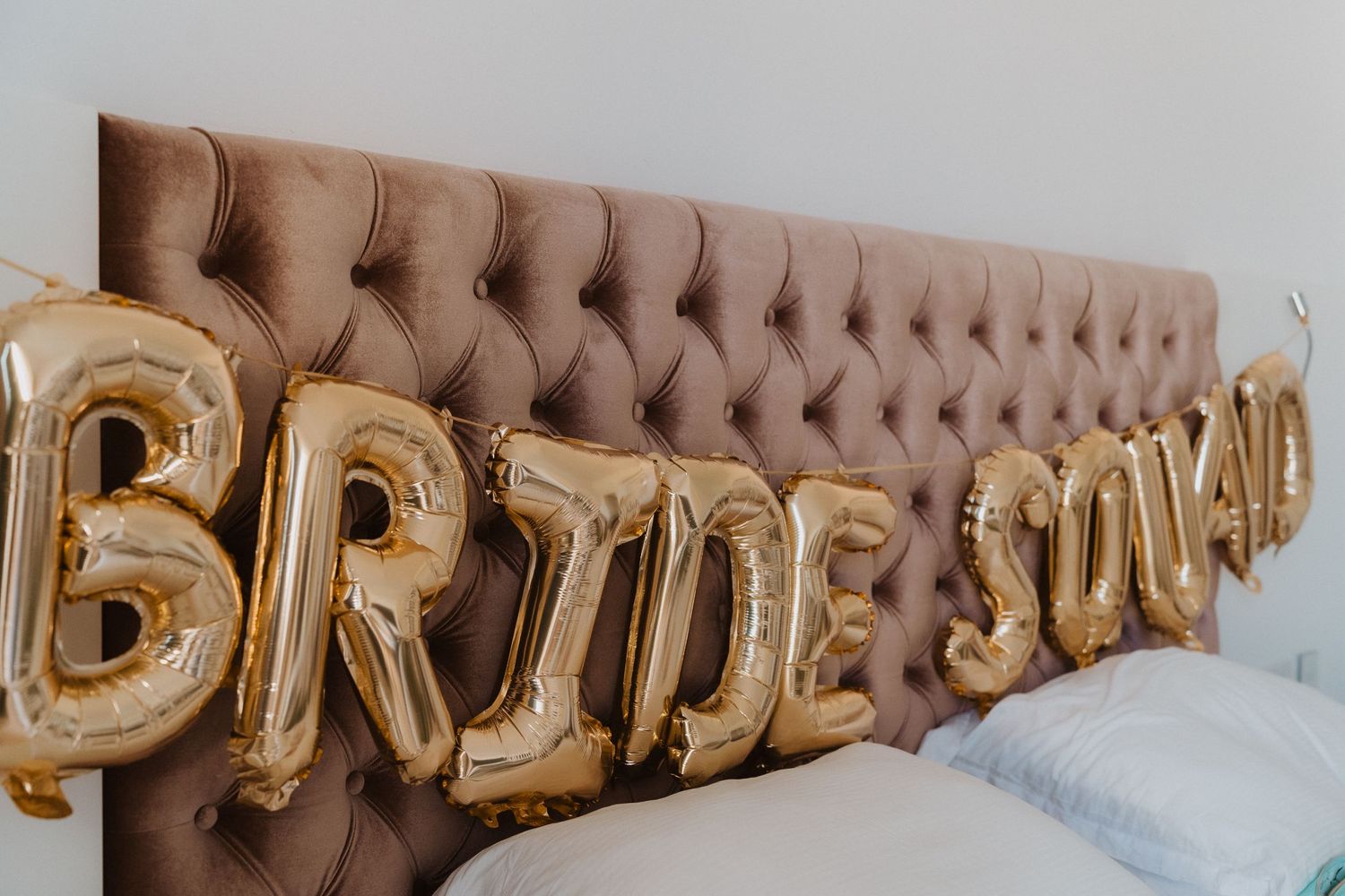 Gold foil 'BRIDE SQUAD' balloons displayed against a tufted brown velvet headboard.
