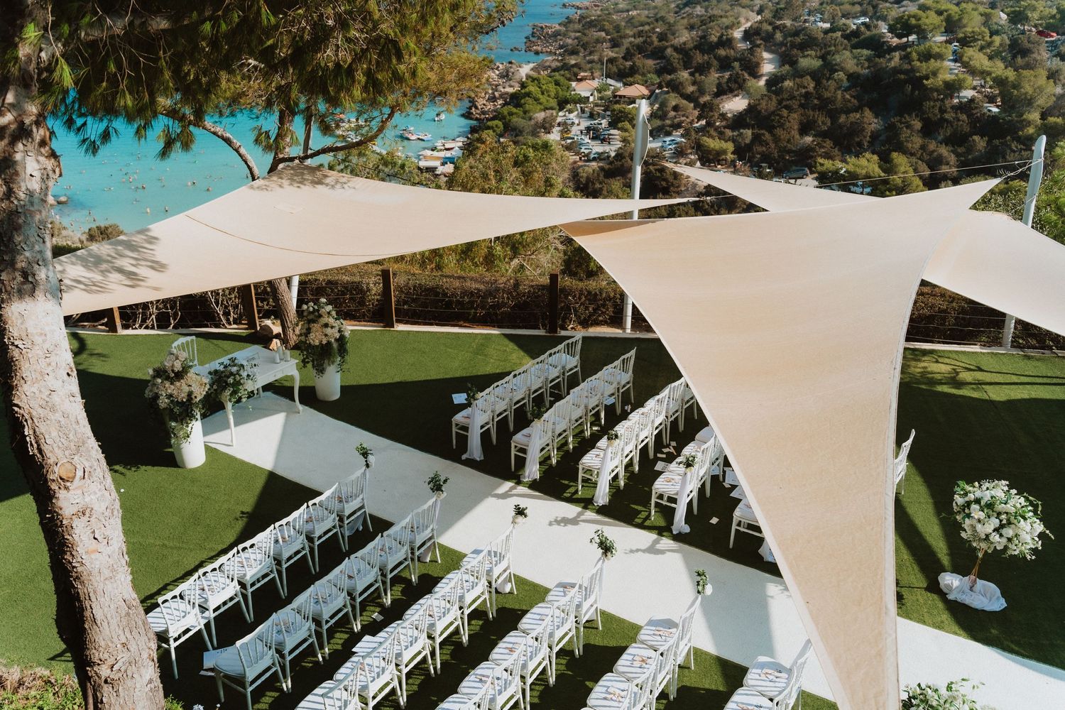Outdoor wedding ceremony setup with white chairs and elegant sail cloth canopies providing shade on a manicured lawn.