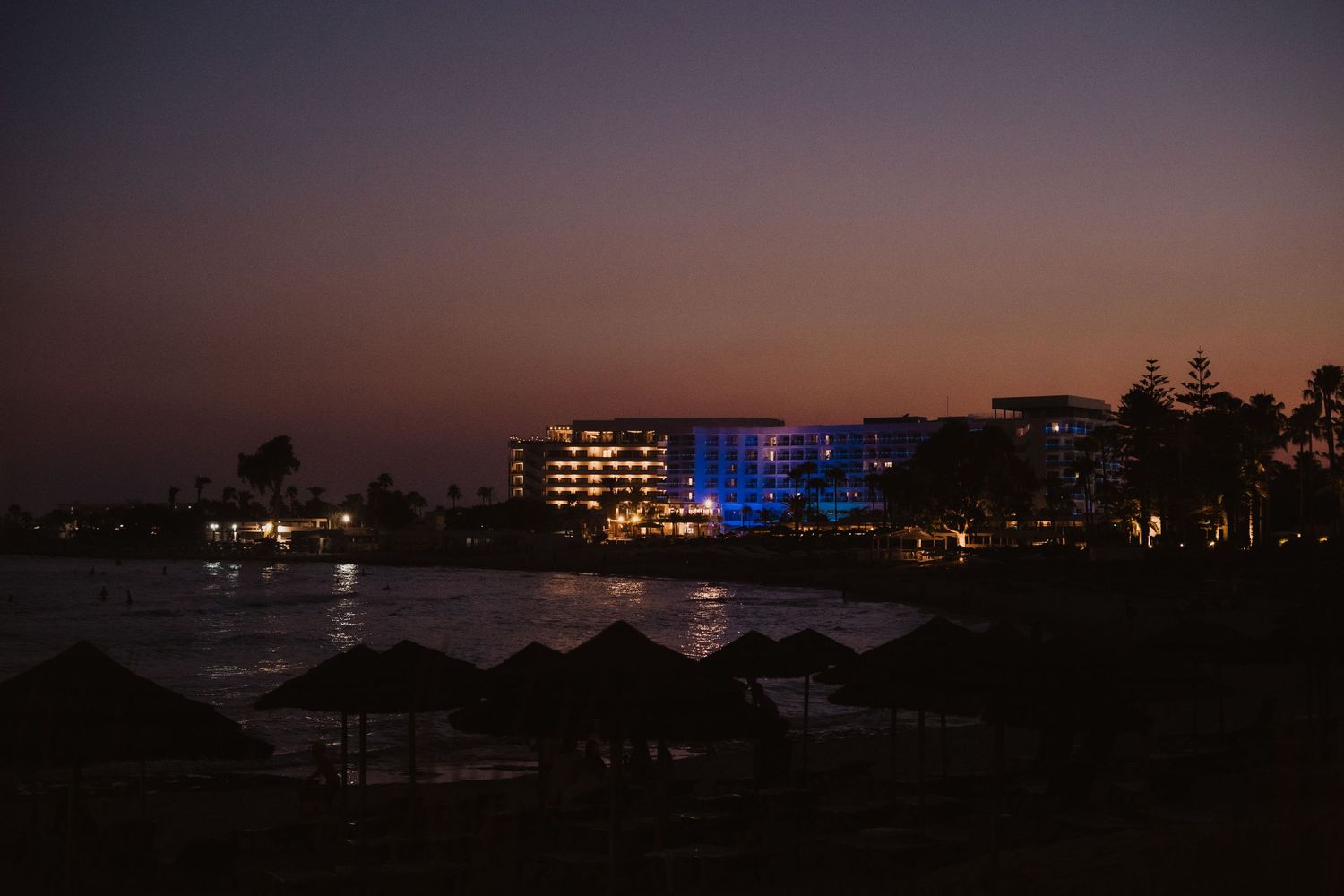 Beachfront resort buildings illuminated at twilight with palm trees and umbrellas silhouetted.