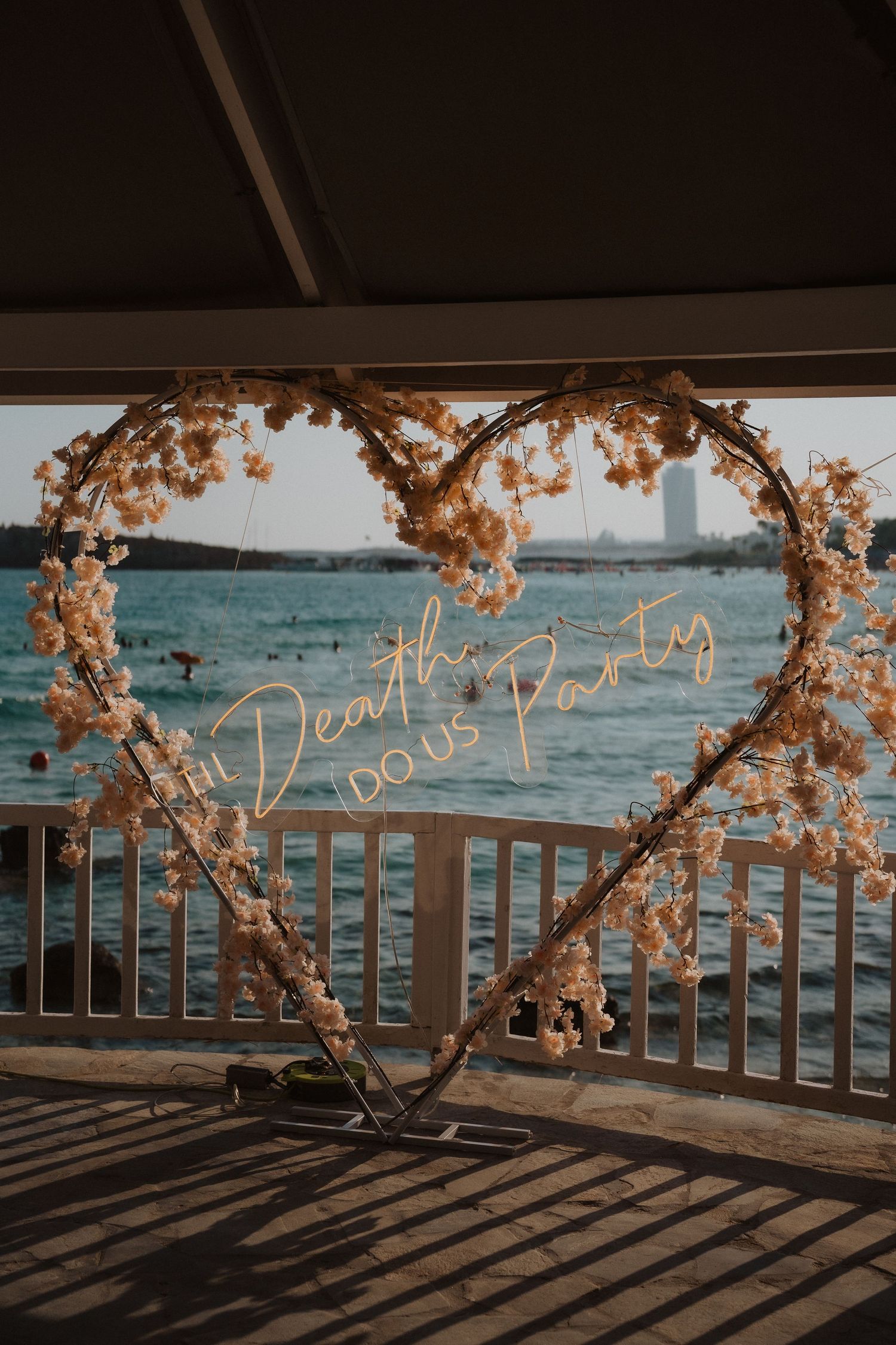 Heart-shaped floral arch with neon 'Death Do Us Part' sign overlooking waterfront at sunset during wedding ceremony.
