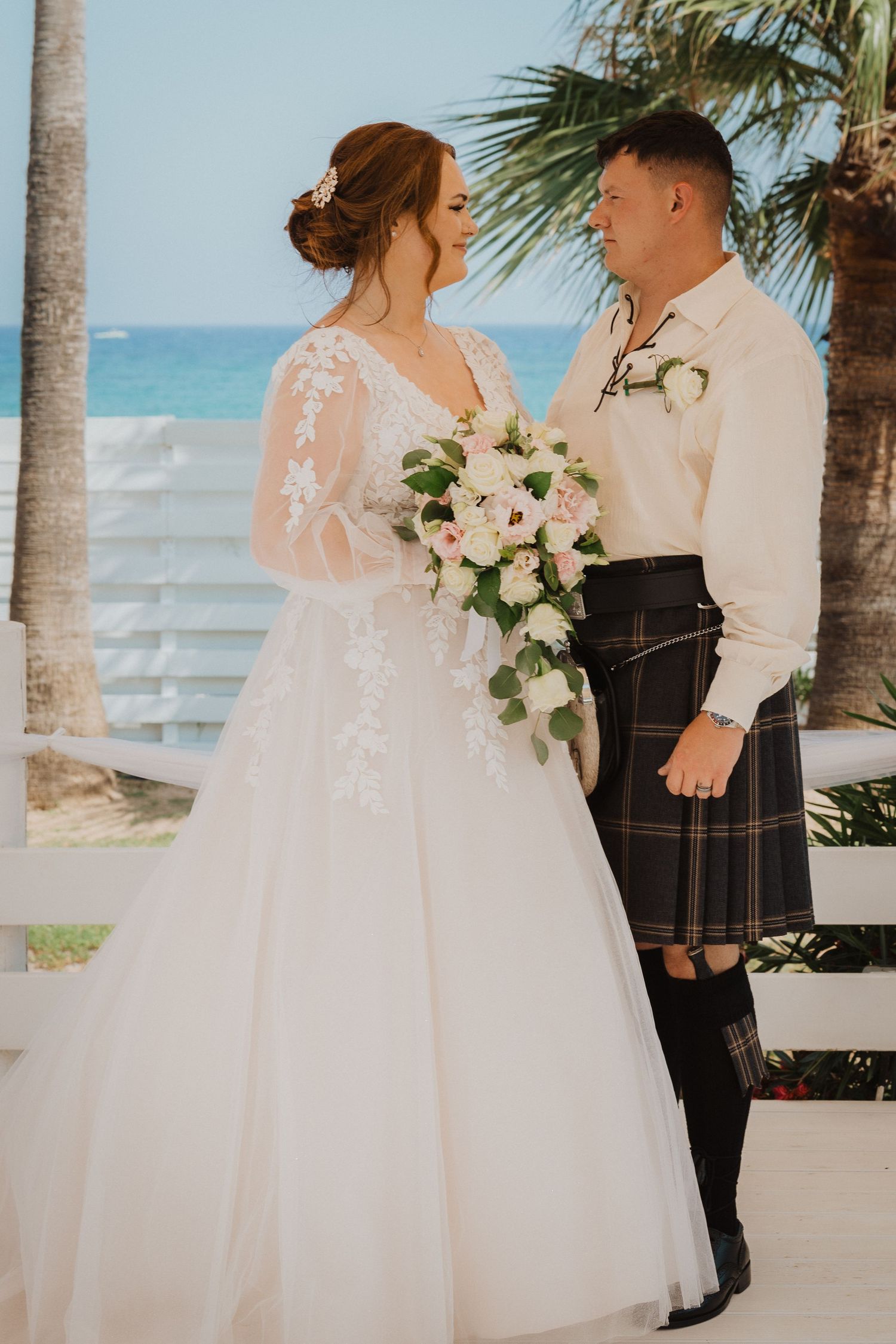 Elegant beach wedding portrait with groom in traditional Scottish kilt and bride holding white floral bouquet.