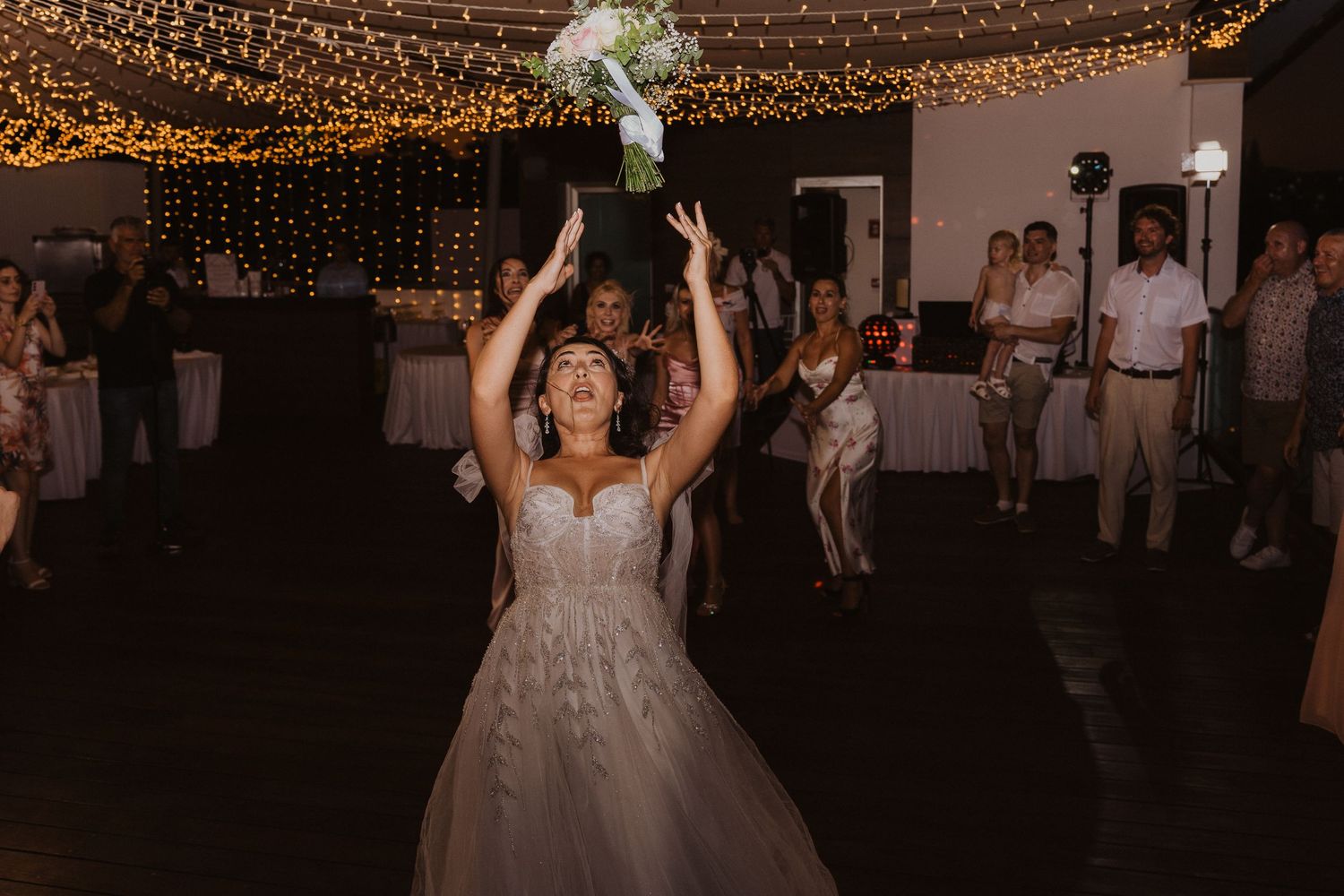 Bride tosses her bouquet during traditional wedding reception moment under sparkling lights.
