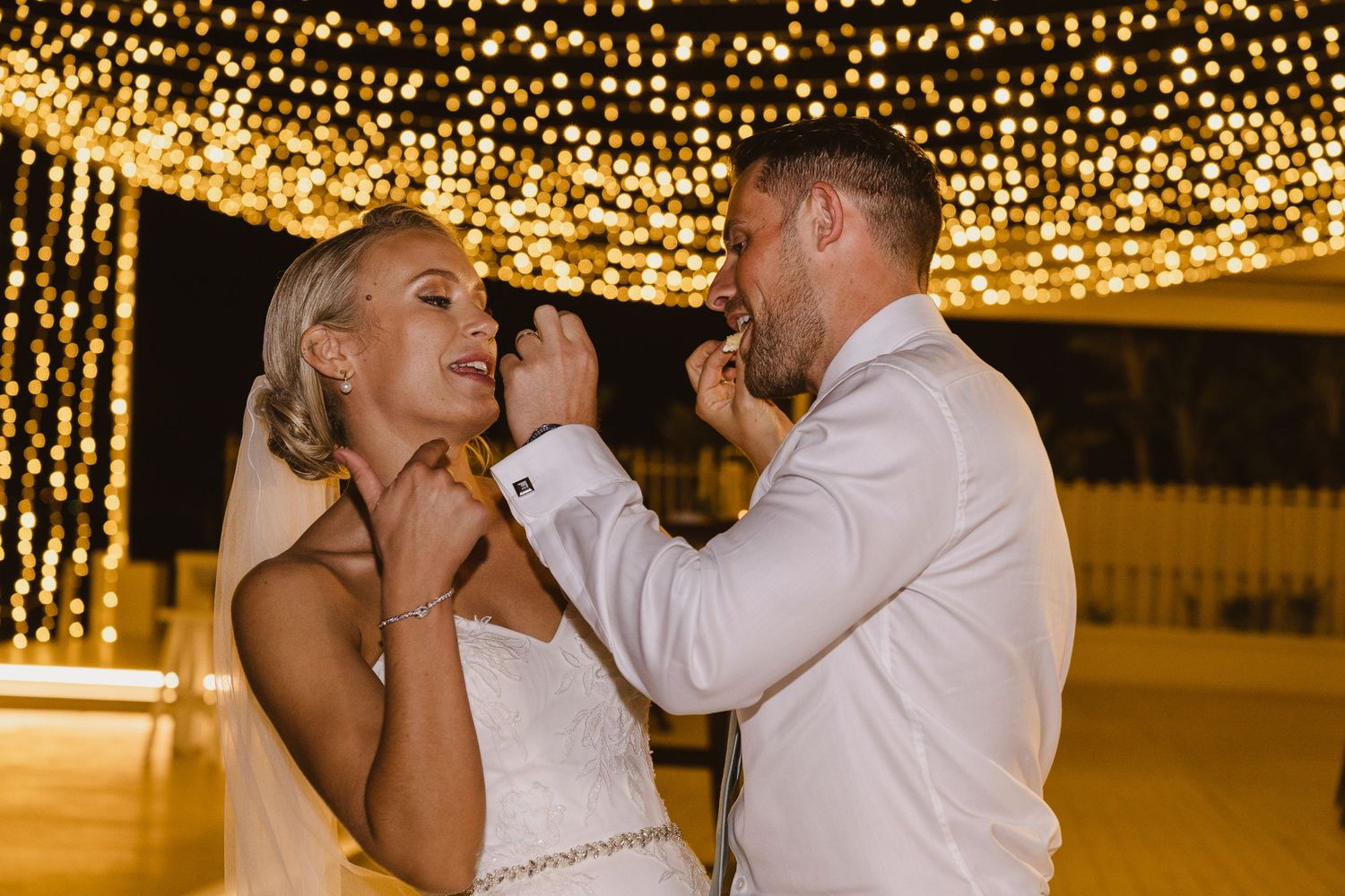 Wedding couple laughs while feeding each other cake beneath glowing string lights.