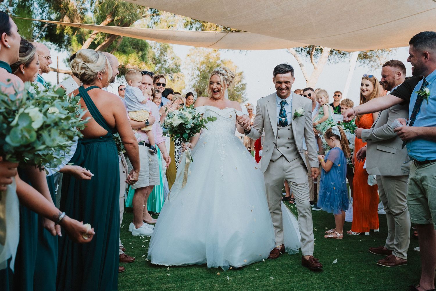 Joyful newlyweds walk down aisle while wedding guests celebrate with flower petals and cheers.