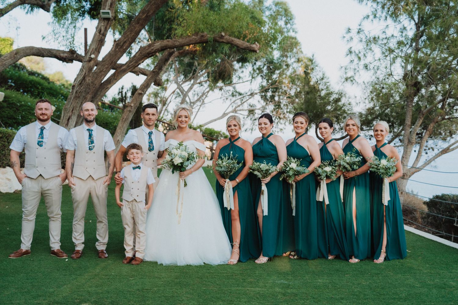 Outdoor wedding party in coordinated emerald green dresses and tan suits pose together on a grassy lawn by the ocean.