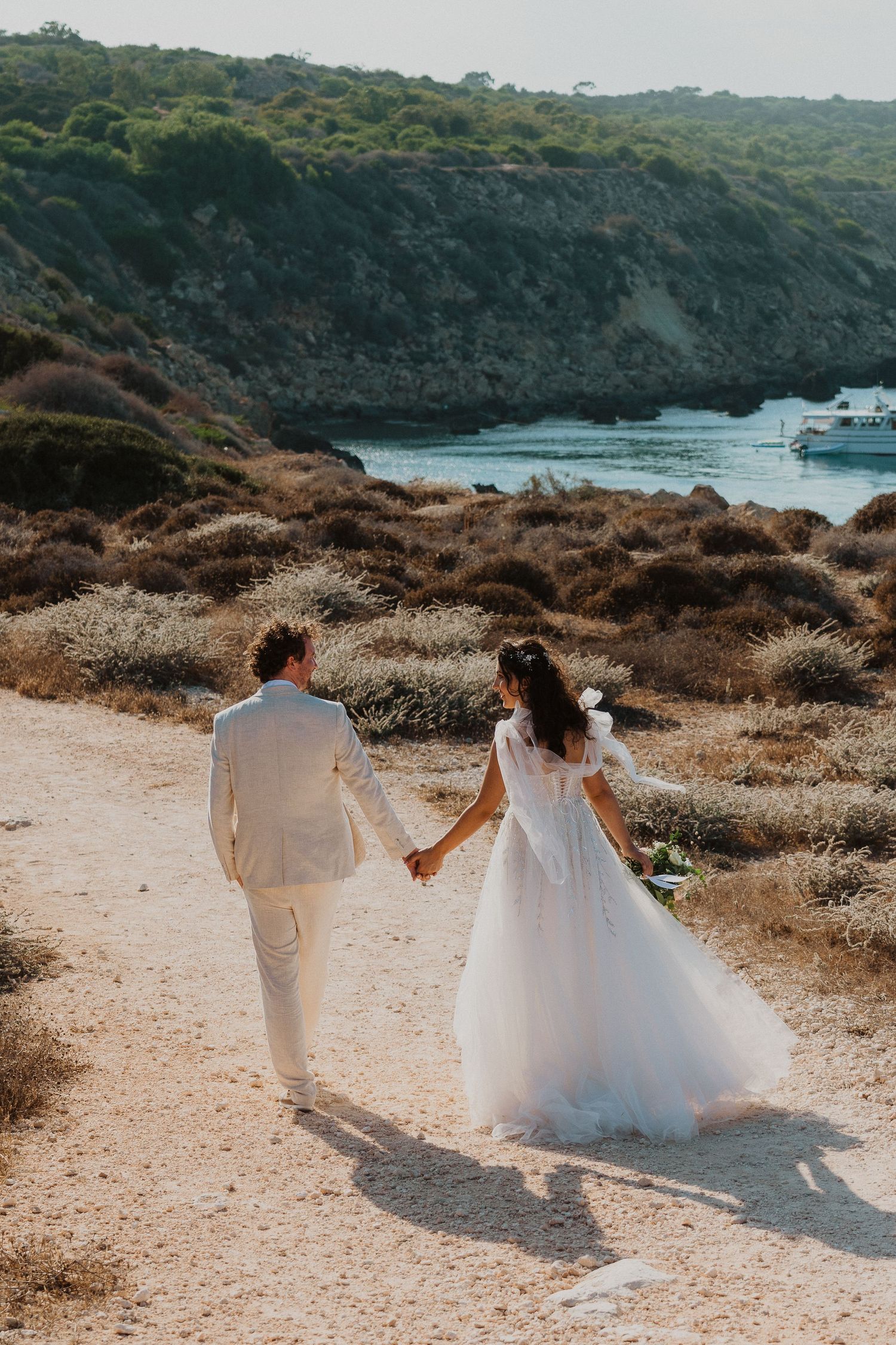 A couple in wedding attire walking hand in hand along a coastal path with a yacht anchored in the turquoise bay below.