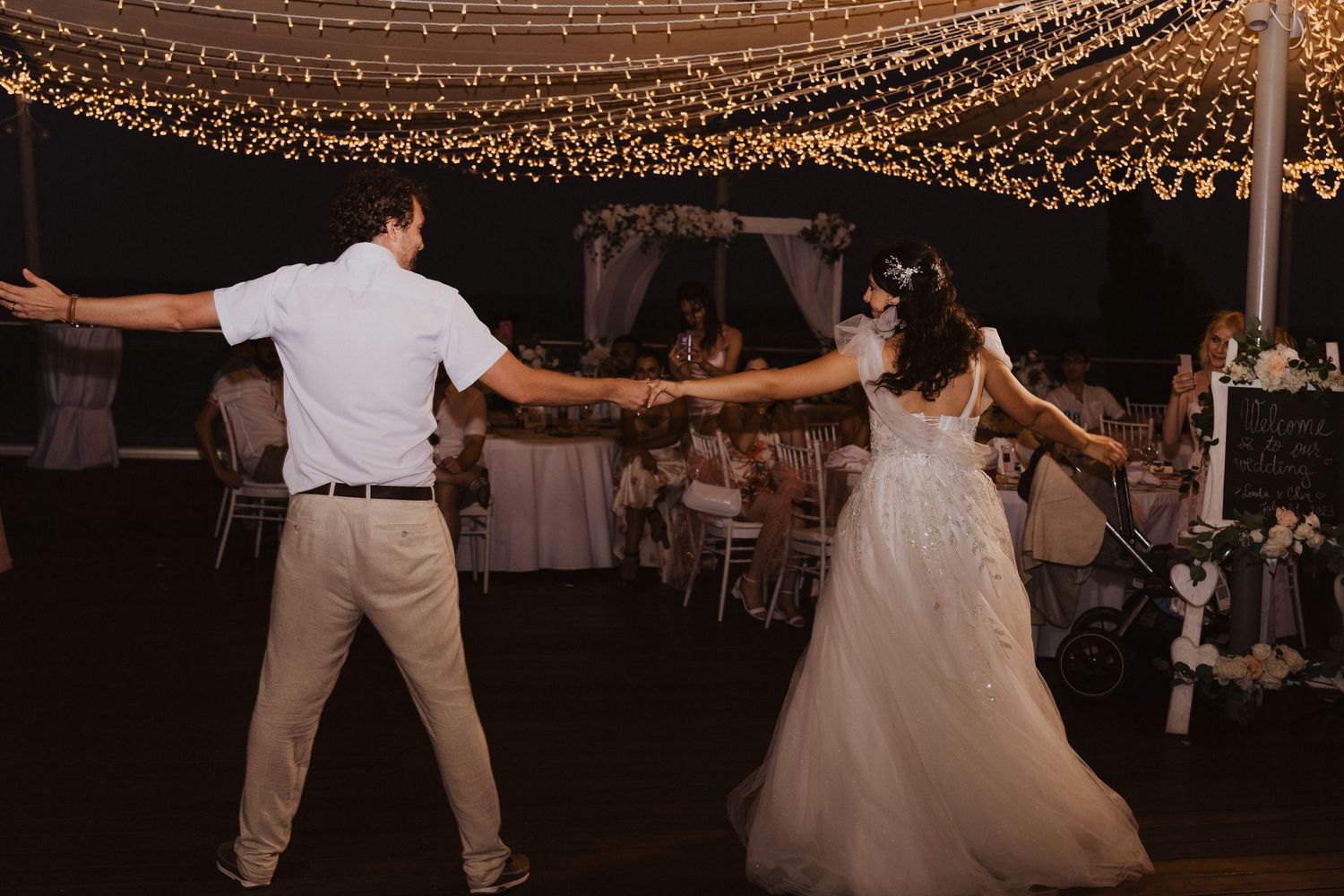 A couple performs an energetic dance move during their wedding reception under twinkling lights.