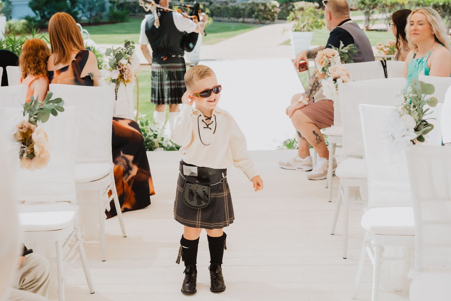 Small child in formal attire and sunglasses walking confidently down the wedding aisle.