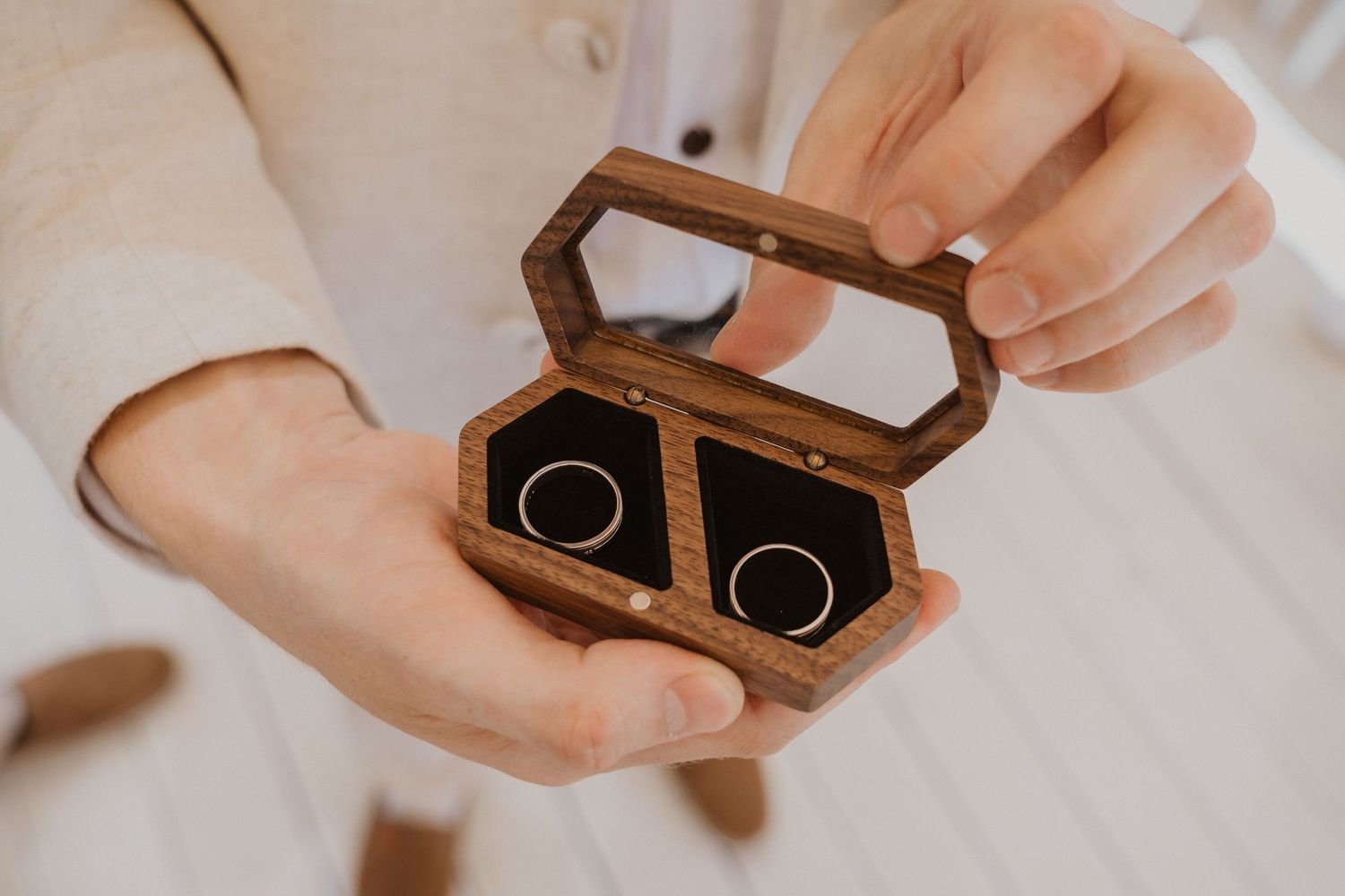 Hands holding open a small wooden ring box with two wedding bands nestled inside.