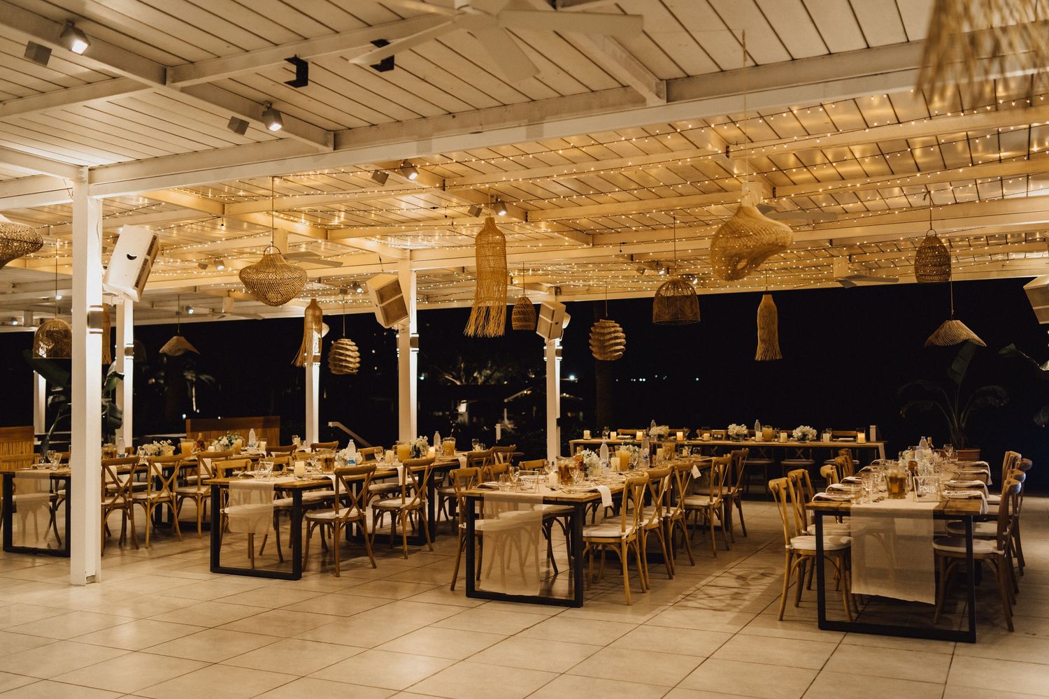 Large wedding reception hall with wooden tables and chairs illuminated by warm string lights.