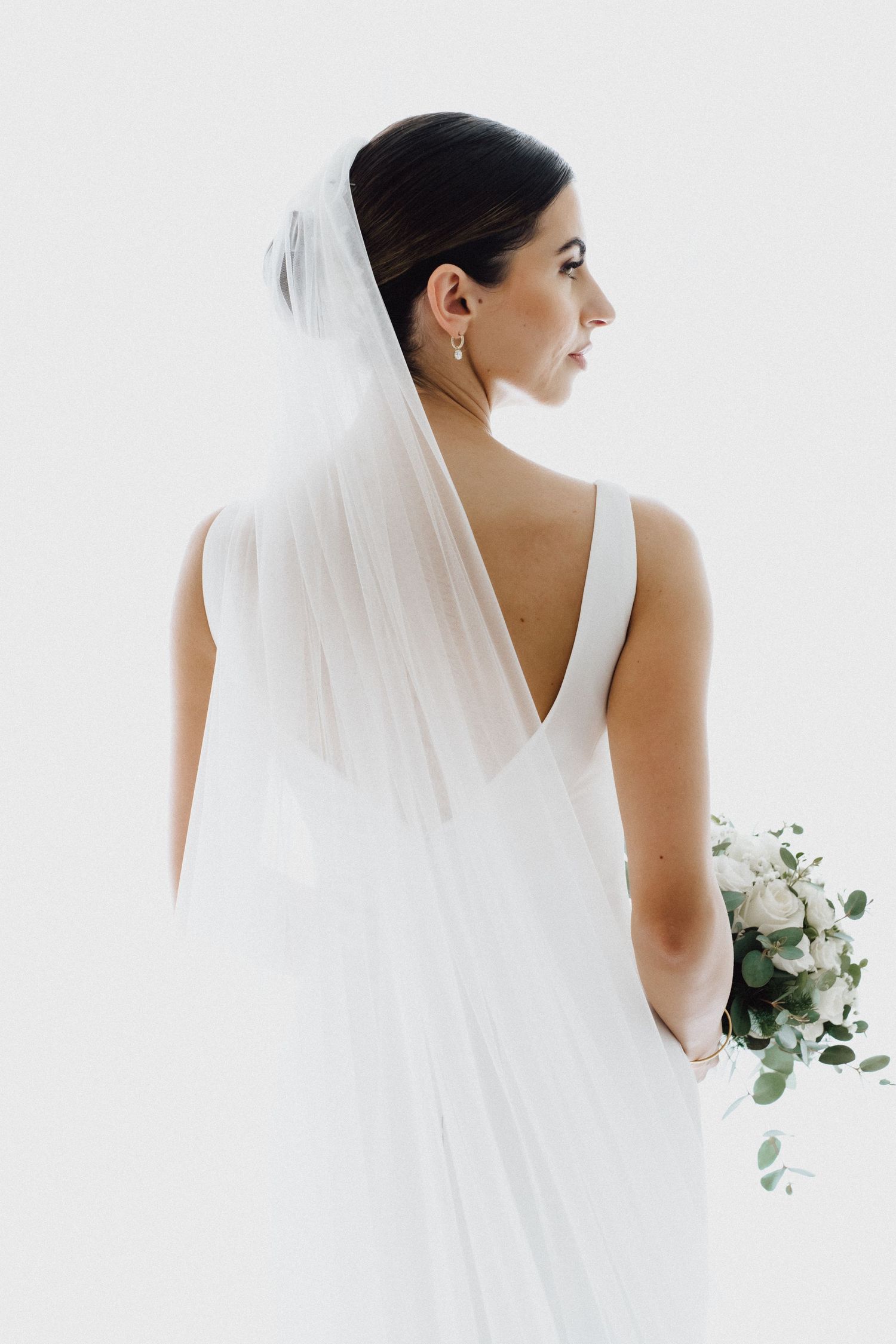 Side profile of bride in elegant white V-back wedding dress holding delicate floral bouquet.