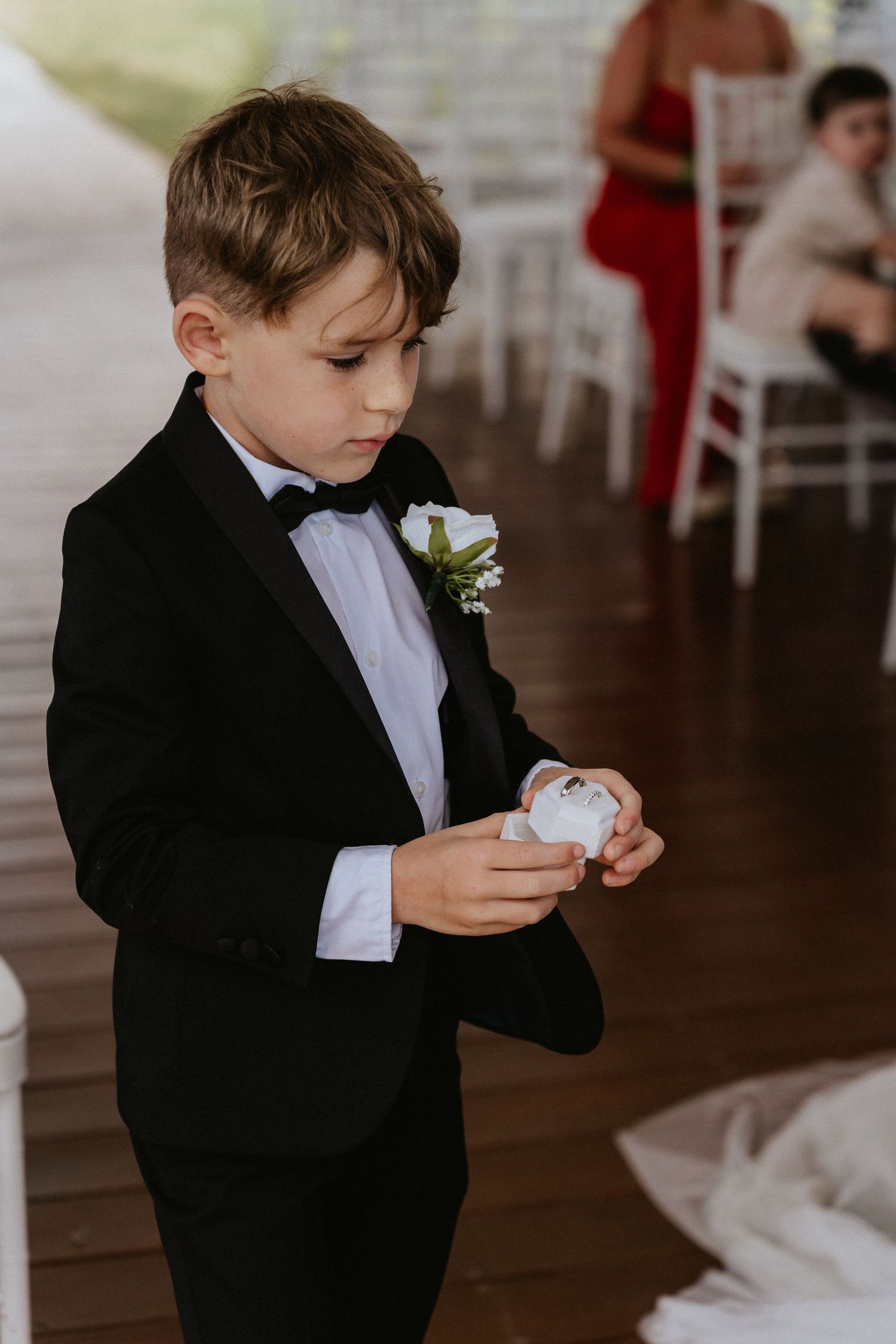 Young ring bearer in black tuxedo holding something precious during wedding ceremony.