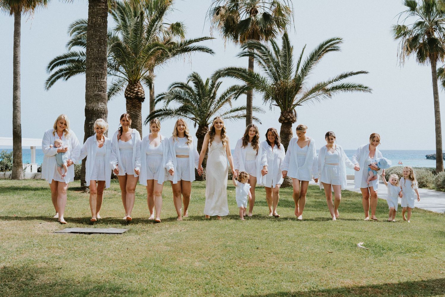 Wedding party in all-white attire poses under palm trees at a beachfront resort.