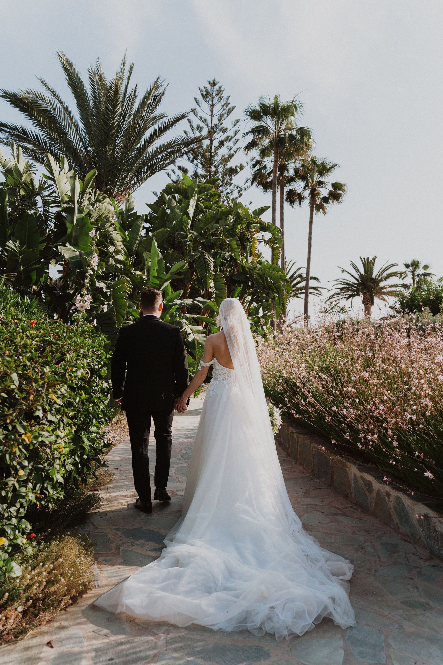 Stunning wedding photo of couple walking through palm-lined garden path with flowing white dress train.
