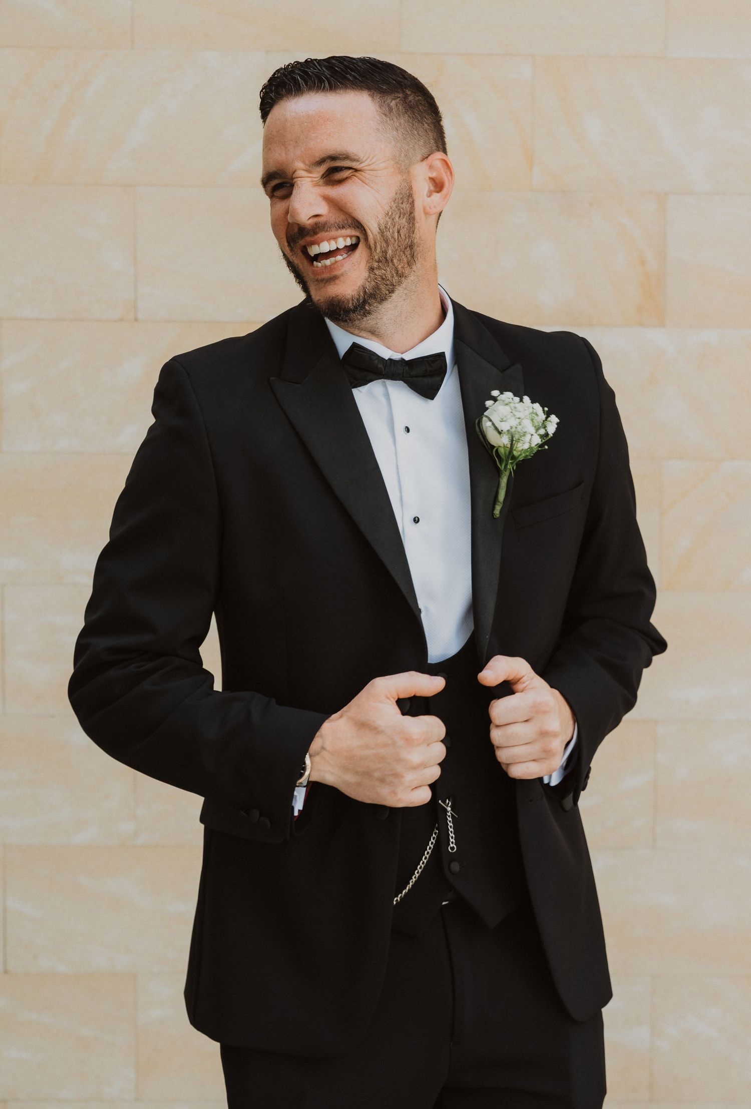 A groom in a black tuxedo and bow tie laughs joyfully while adjusting his jacket during a wedding photoshoot.