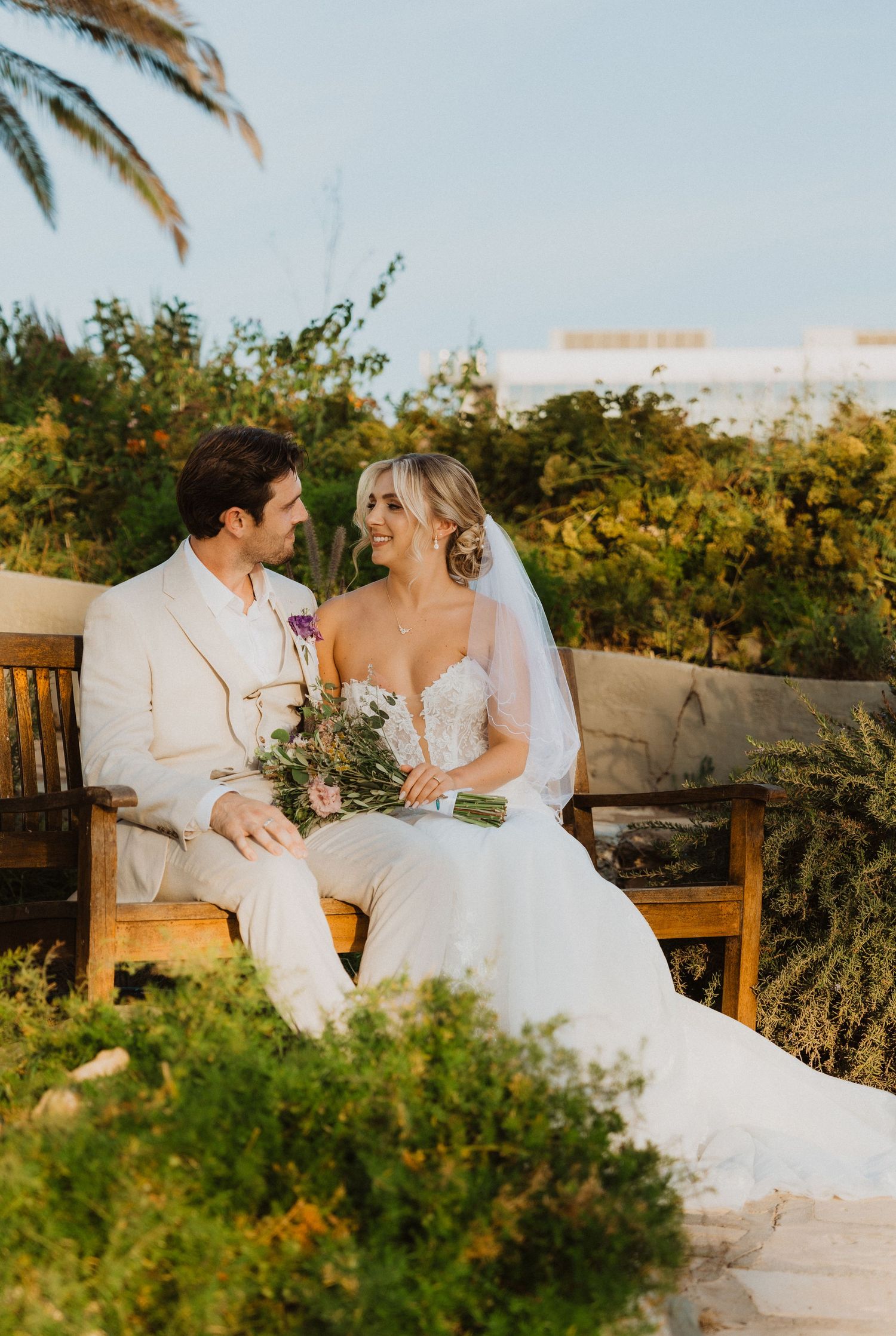 Candid wedding moment captured on garden bench with golden sunlight and tropical foliage.
