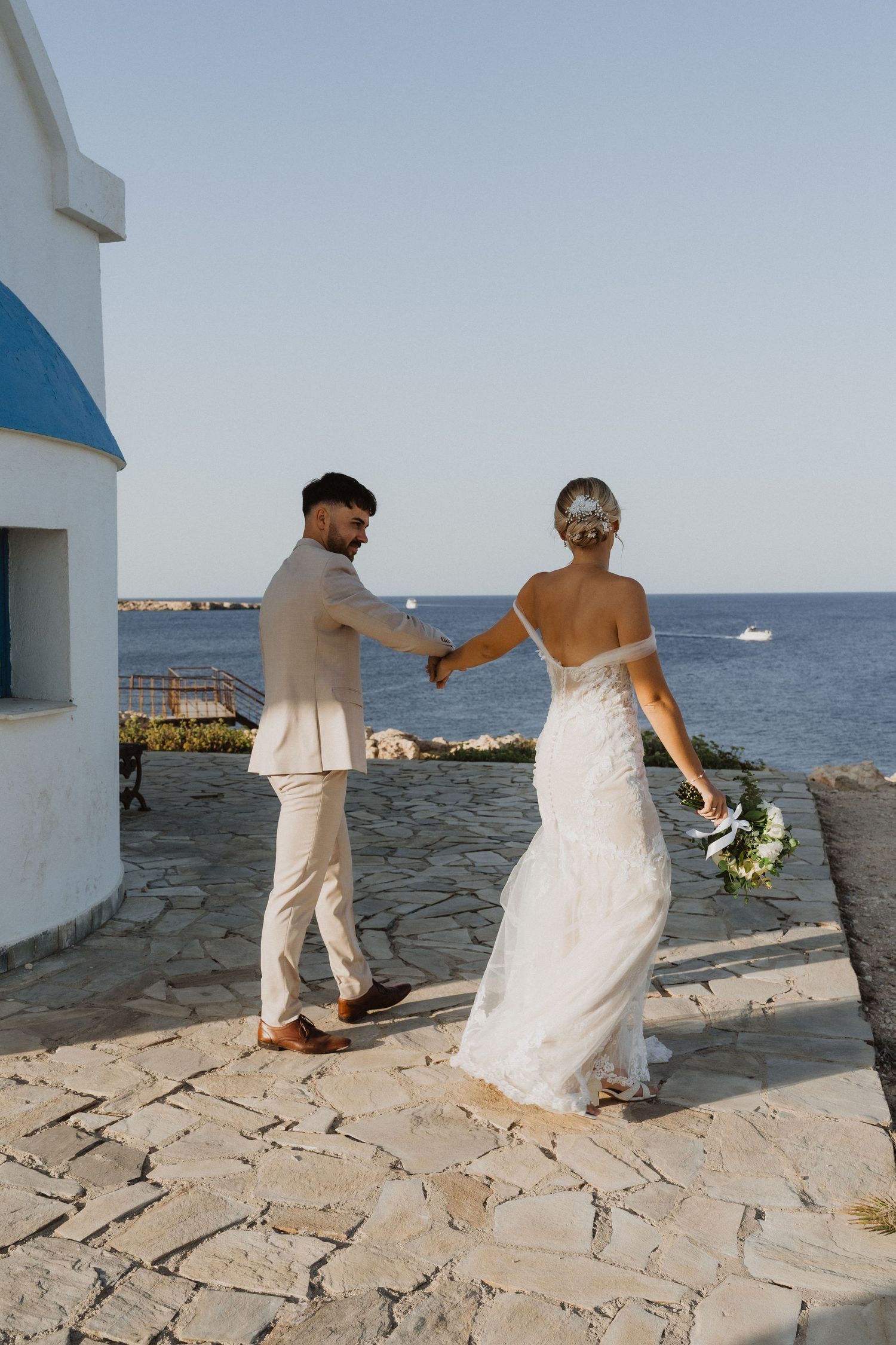 Newlyweds walk along stone terrace overlooking Mediterranean waters.