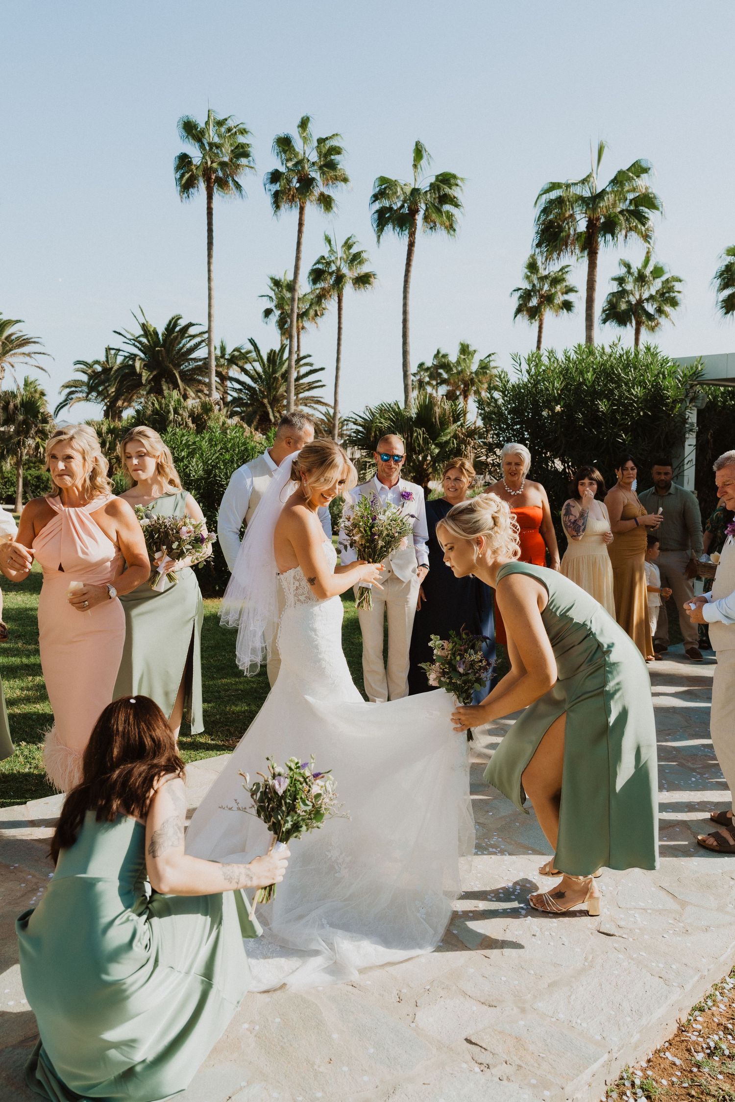 Wedding party celebrating outdoors with palm trees and bridesmaids in sage green dresses at a tropical venue.
