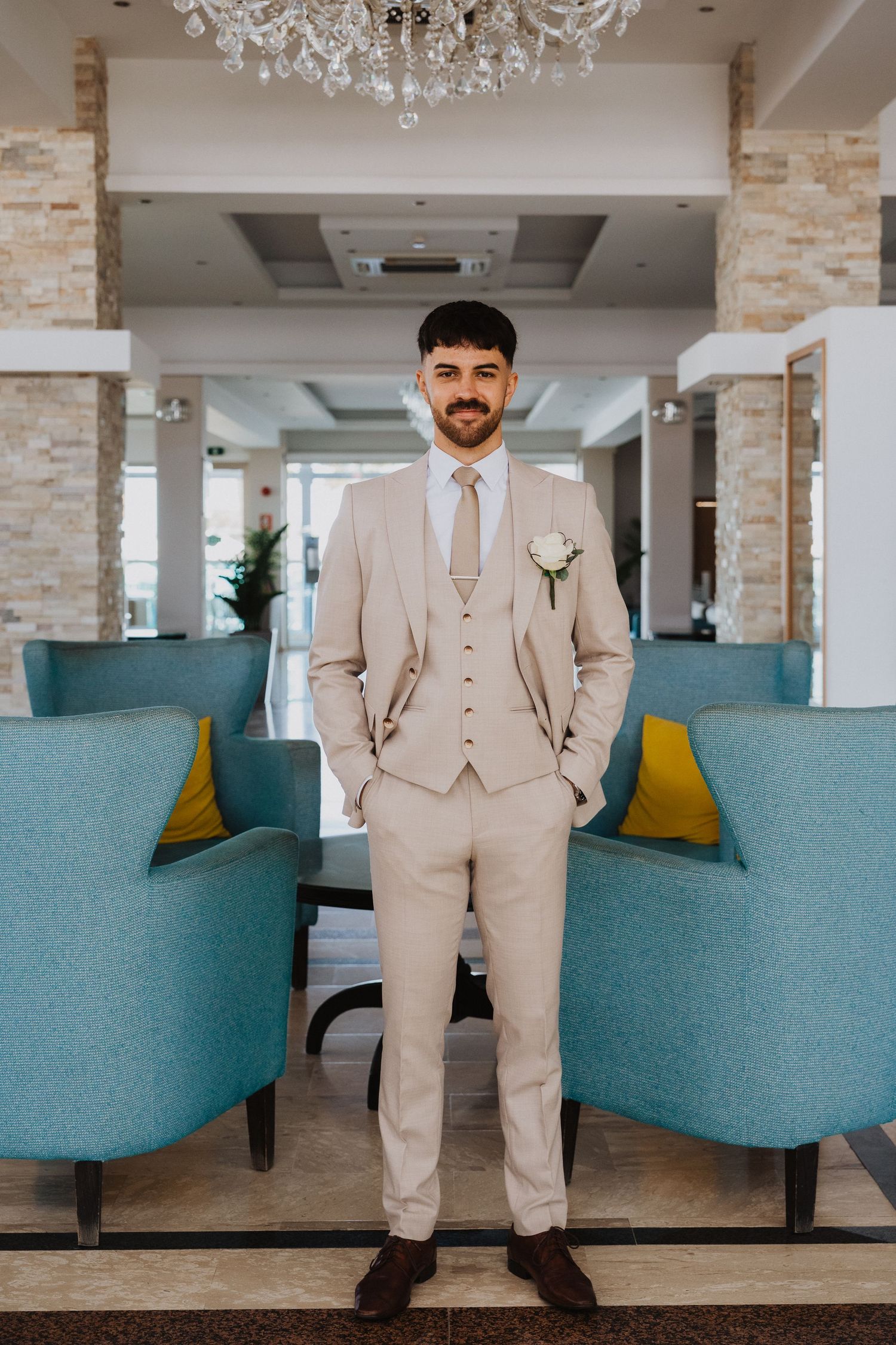 A beige three-piece suit stands in an elegant venue with turquoise chairs and crystal chandeliers.