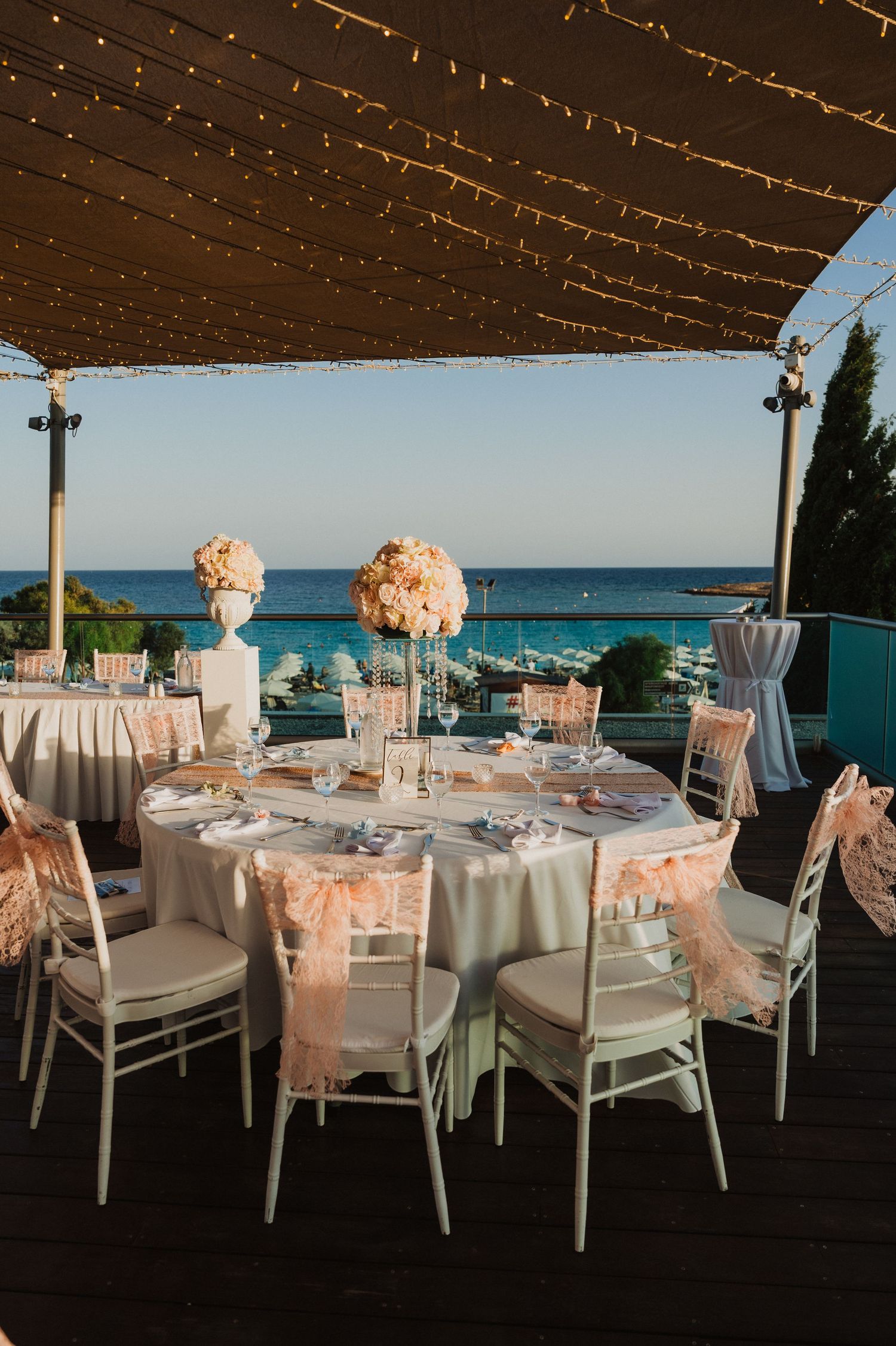 Outdoor wedding reception setup with white tables and chairs overlooking the ocean at sunset.